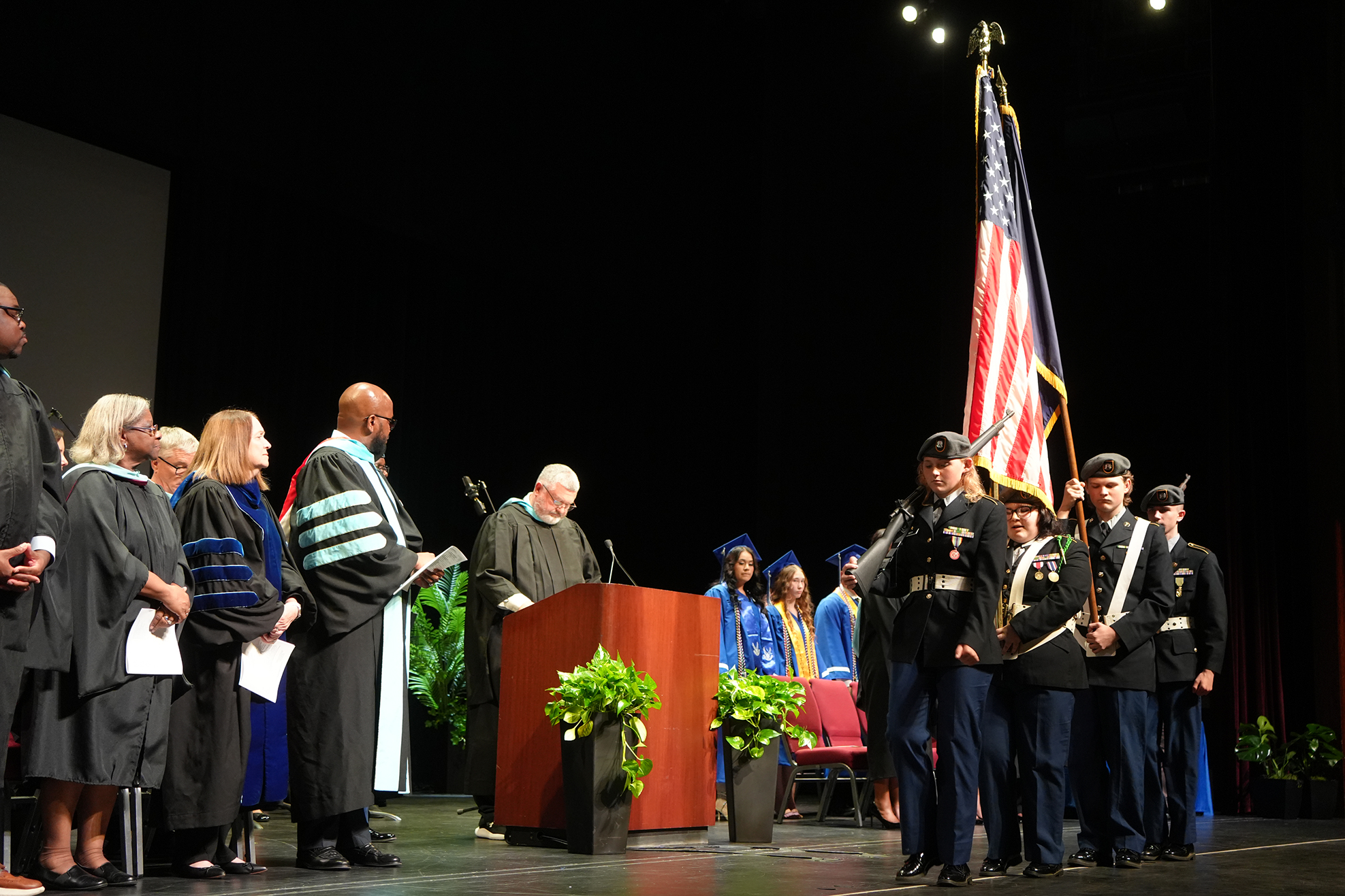 A group of people stand on a stage. A man reads from a podium, and others hold flags.