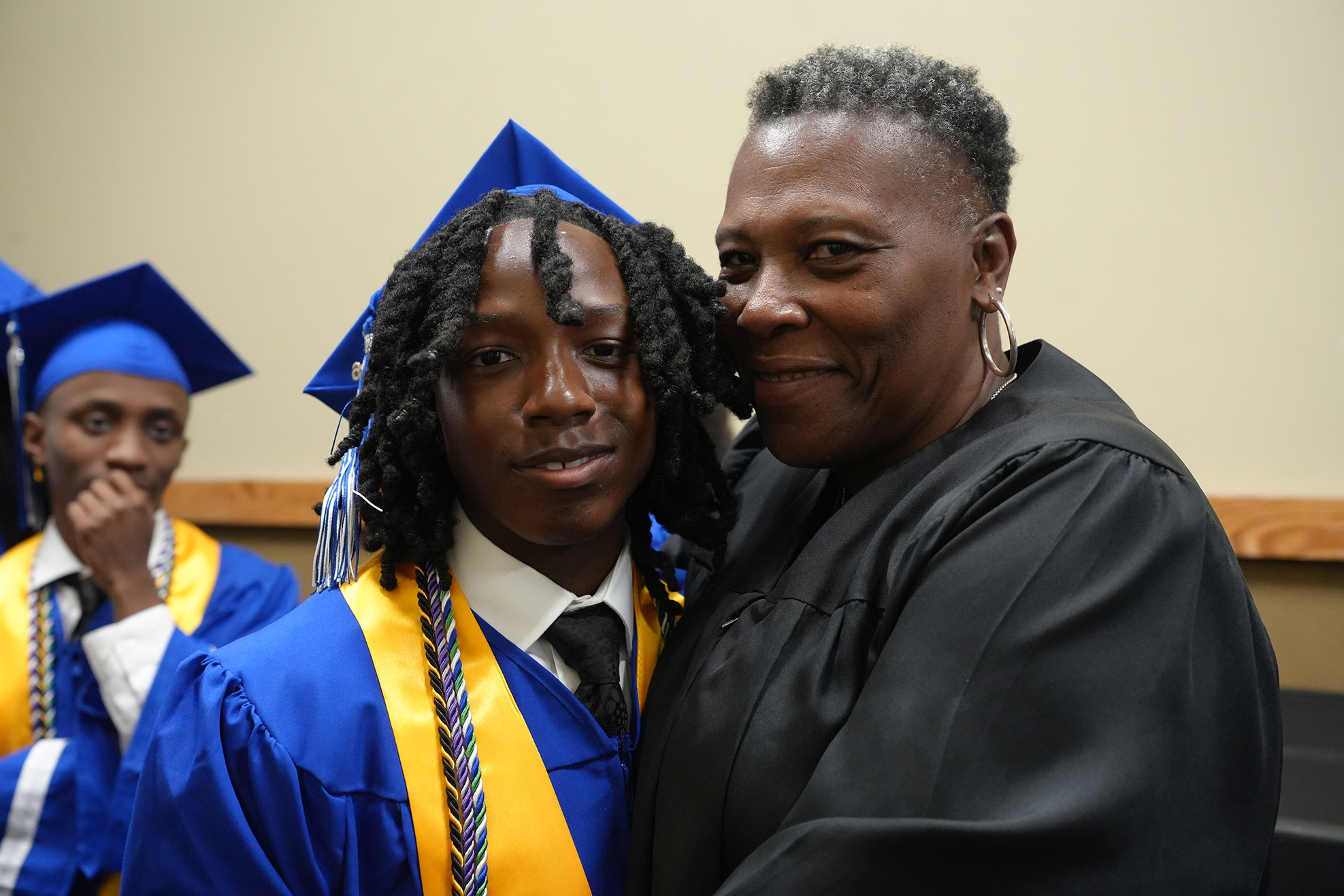 A person in a graduation cap and gown stands with a woman in a black gown, smiling.