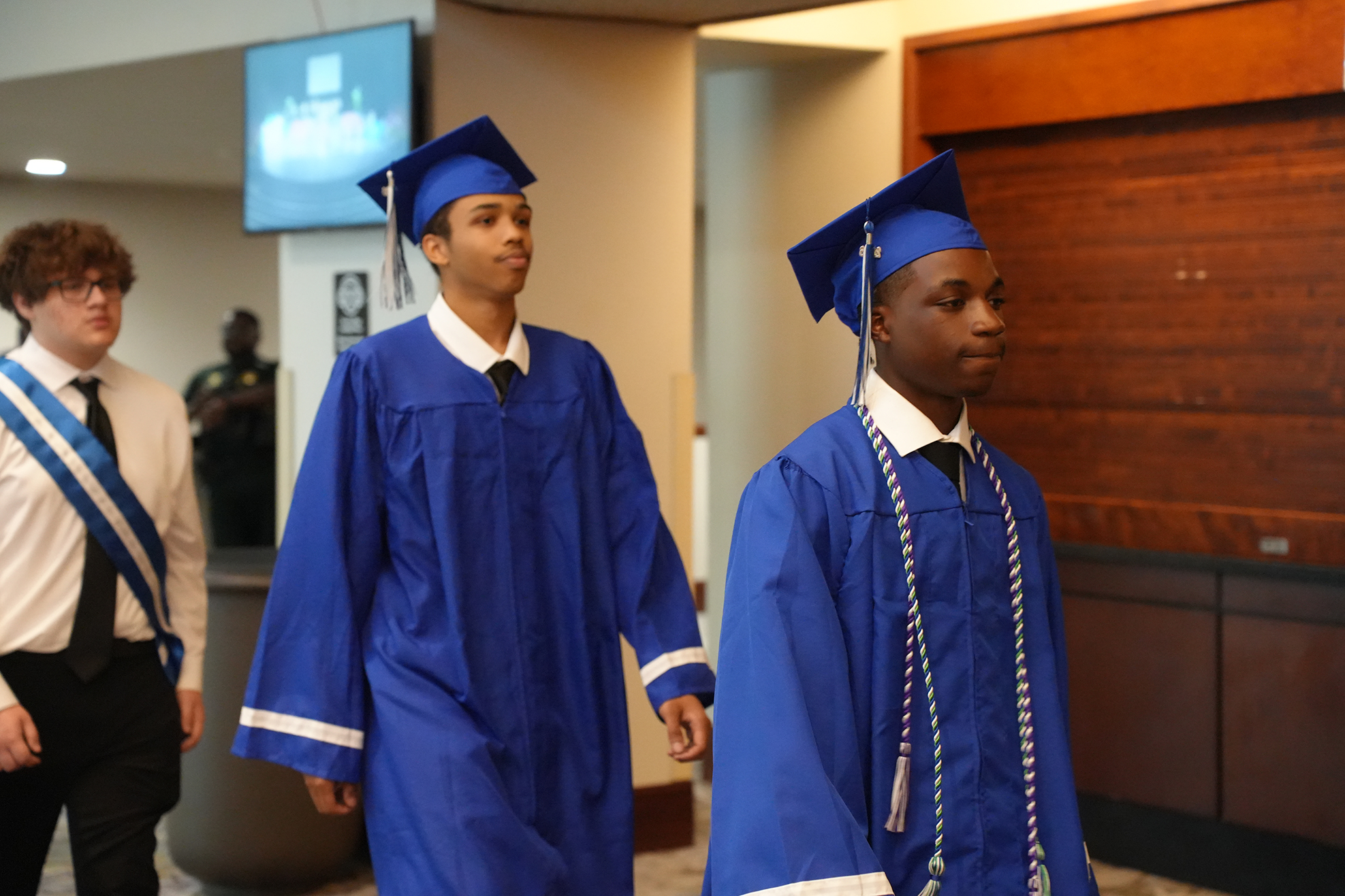 Two young men in blue graduation gowns and caps walk through a hallway. Behind them, a man in a white shirt and tie stands.