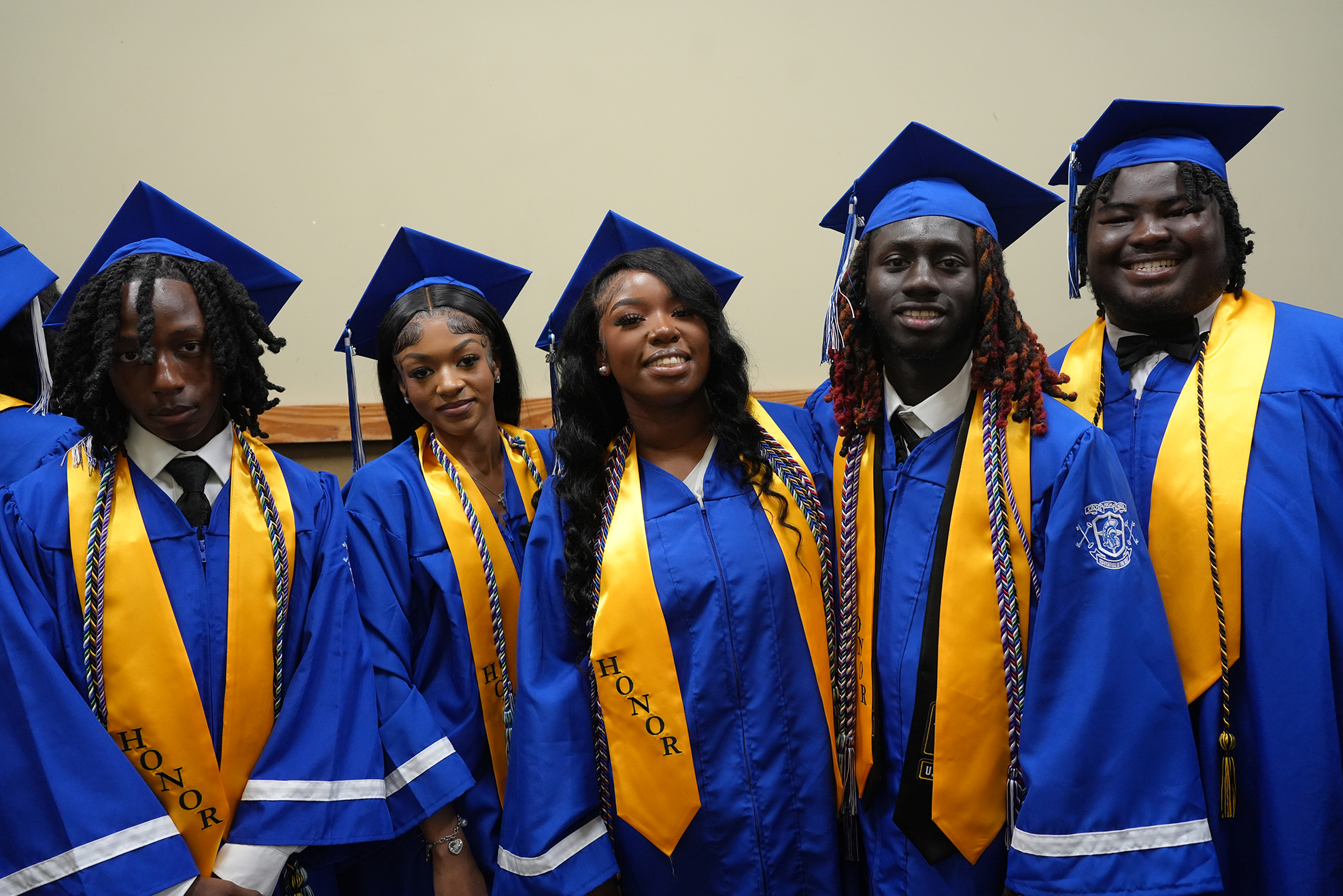 Six individuals in blue gowns with yellow sashes and matching caps stand in a row, smiling for the camera.