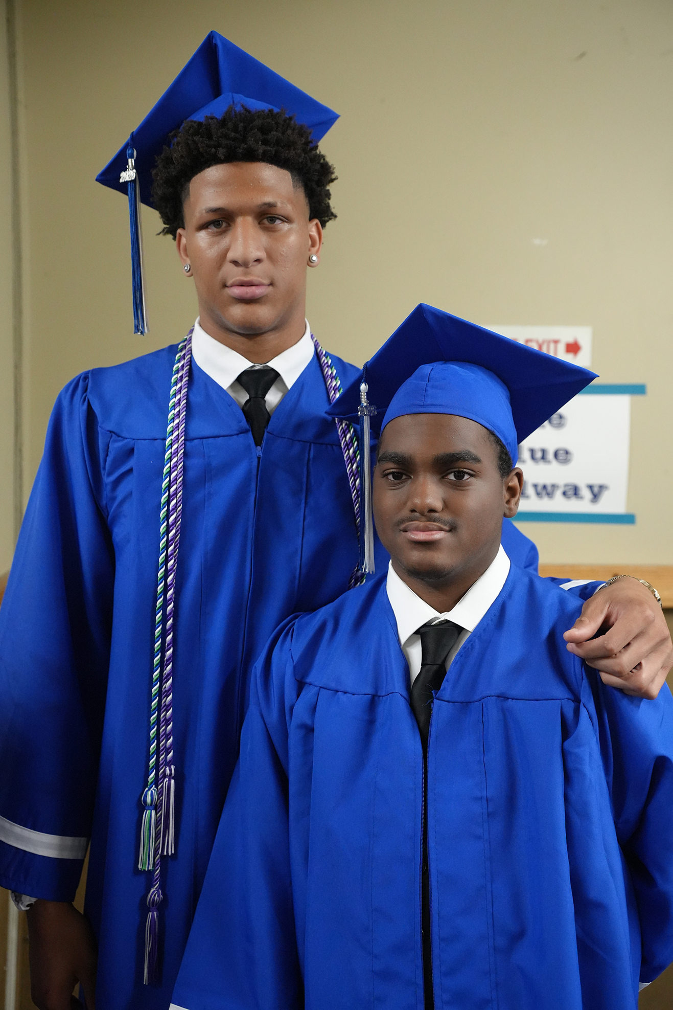 Two young men in blue caps and gowns, standing next to each other, one with his arm around the other.