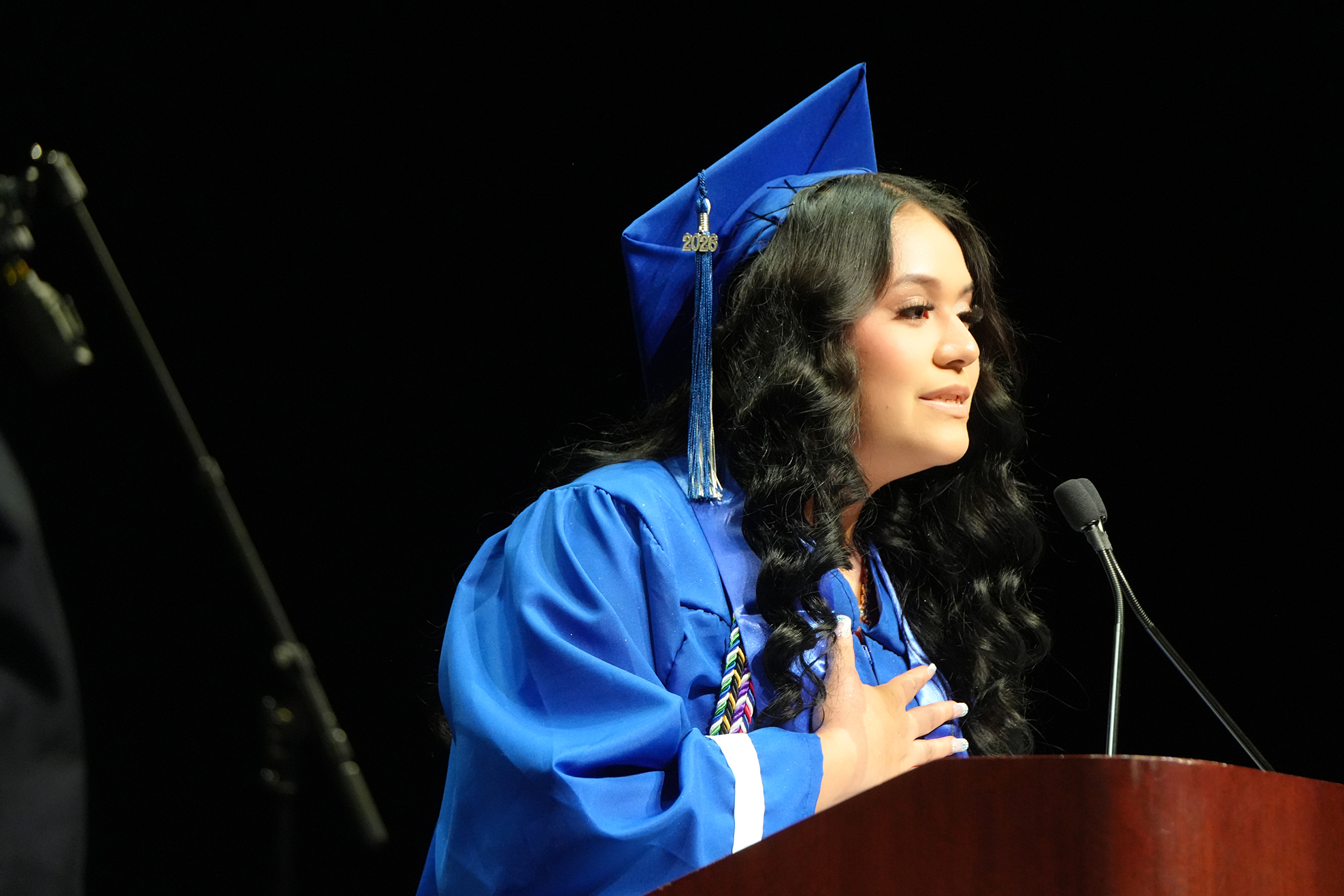 A person in a graduation gown, hat, and necklace speaks into a microphone at a podium.