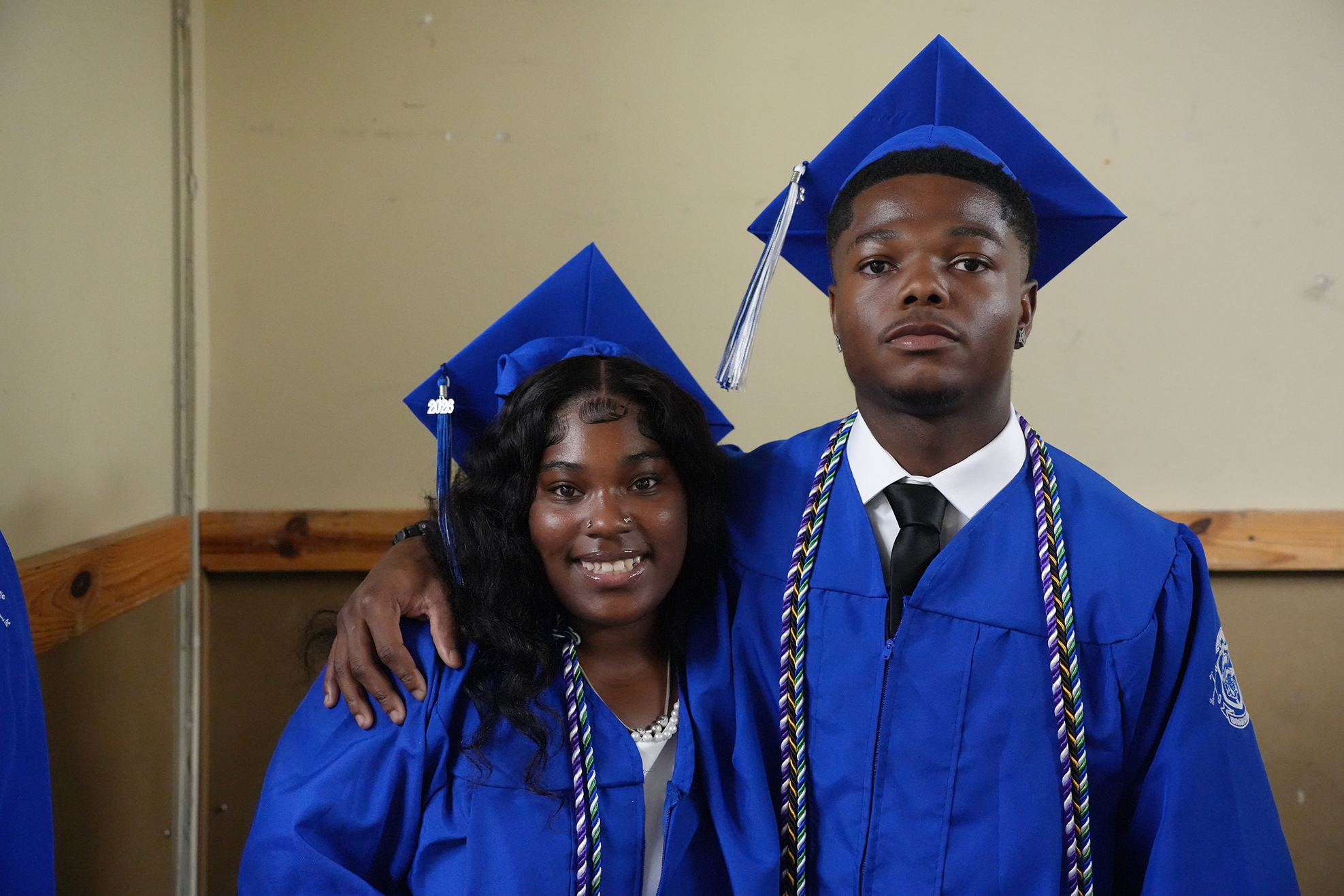 A young man and woman in blue graduation gowns and caps stand close, smiling for the camera.