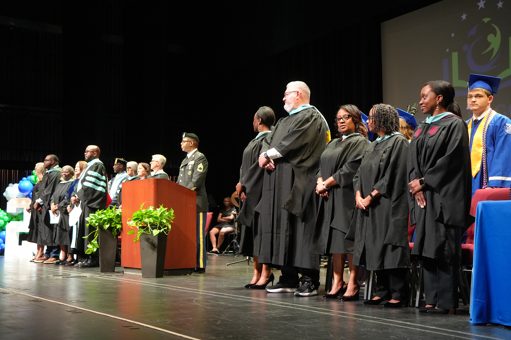Graduates in caps and gowns stand on stage at a ceremony, with a podium and a large screen behind them.