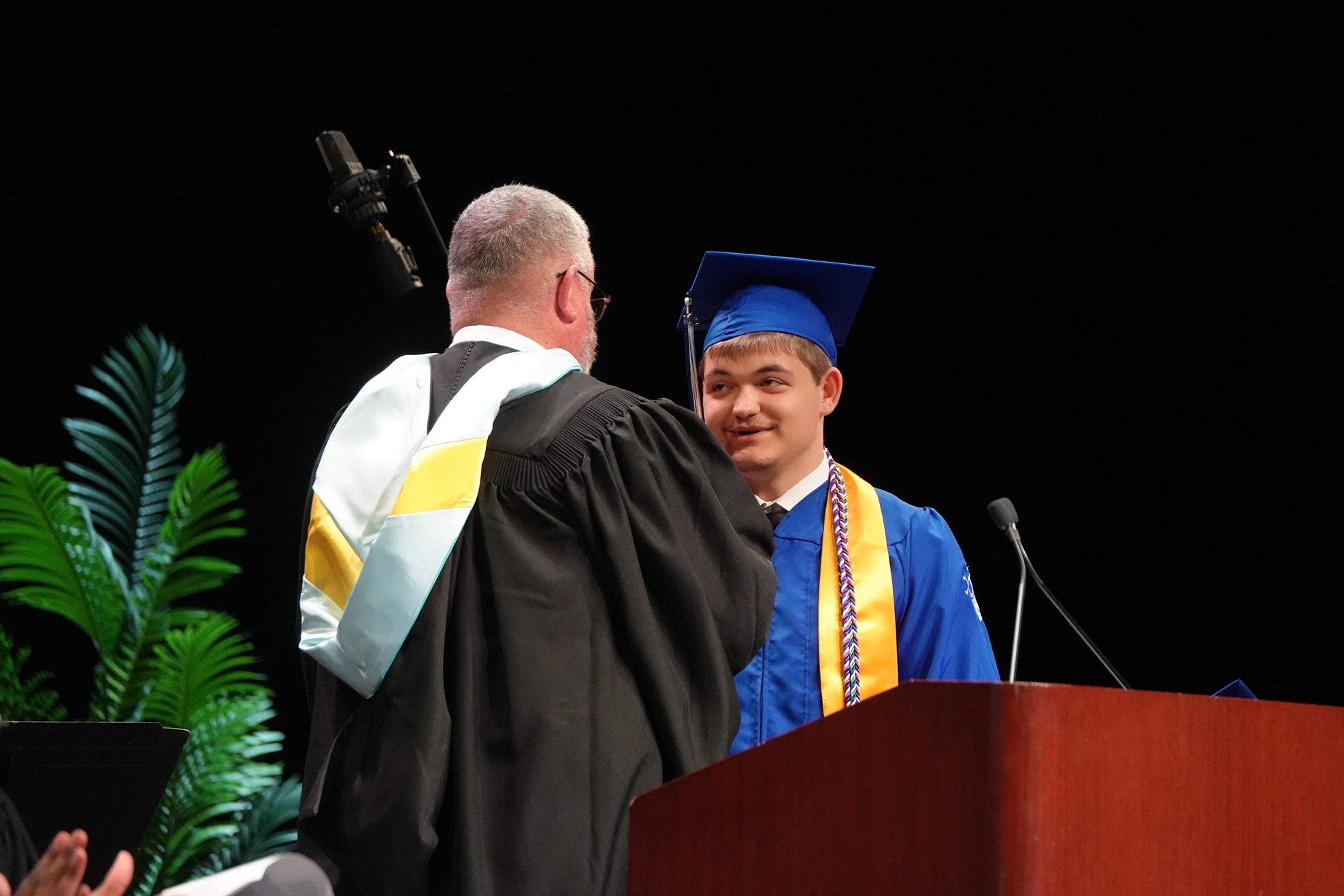 A person in a cap and gown stands by a podium, talking with a person in a black robe.