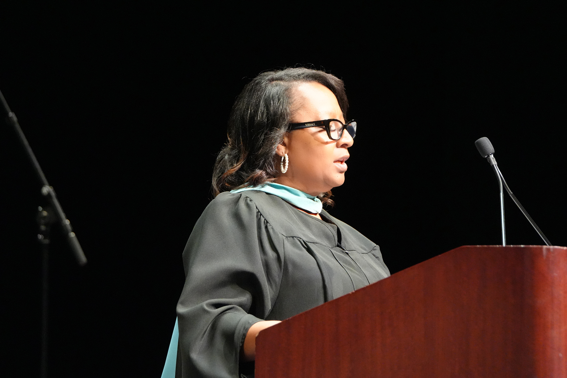 Woman in a black robe and glasses speaks at a podium with a microphone on a dark background.