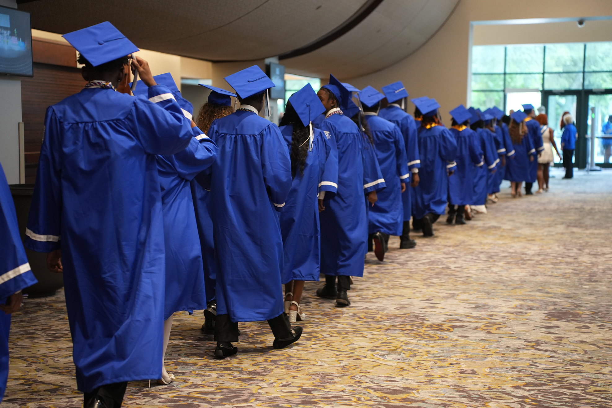 A line of students in blue gowns and hats, possibly waiting in a school hallway with arched ceilings.