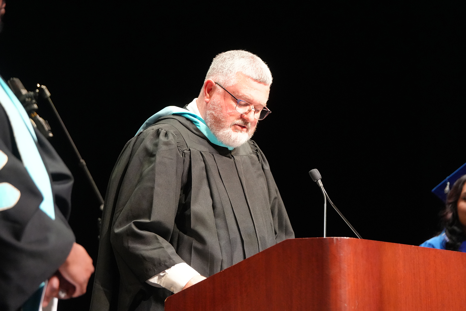 A man in a black academic gown stands at a podium, possibly speaking at a ceremony.