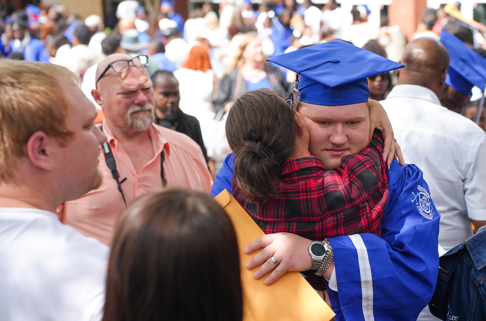 A crowd of people outdoors. A man in a blue graduation cap and gown hugs a woman in a red plaid dress.