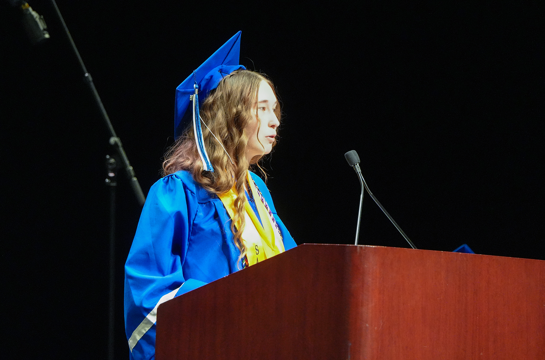 A person in a blue graduation gown stands at a podium with a microphone, looking ahead.