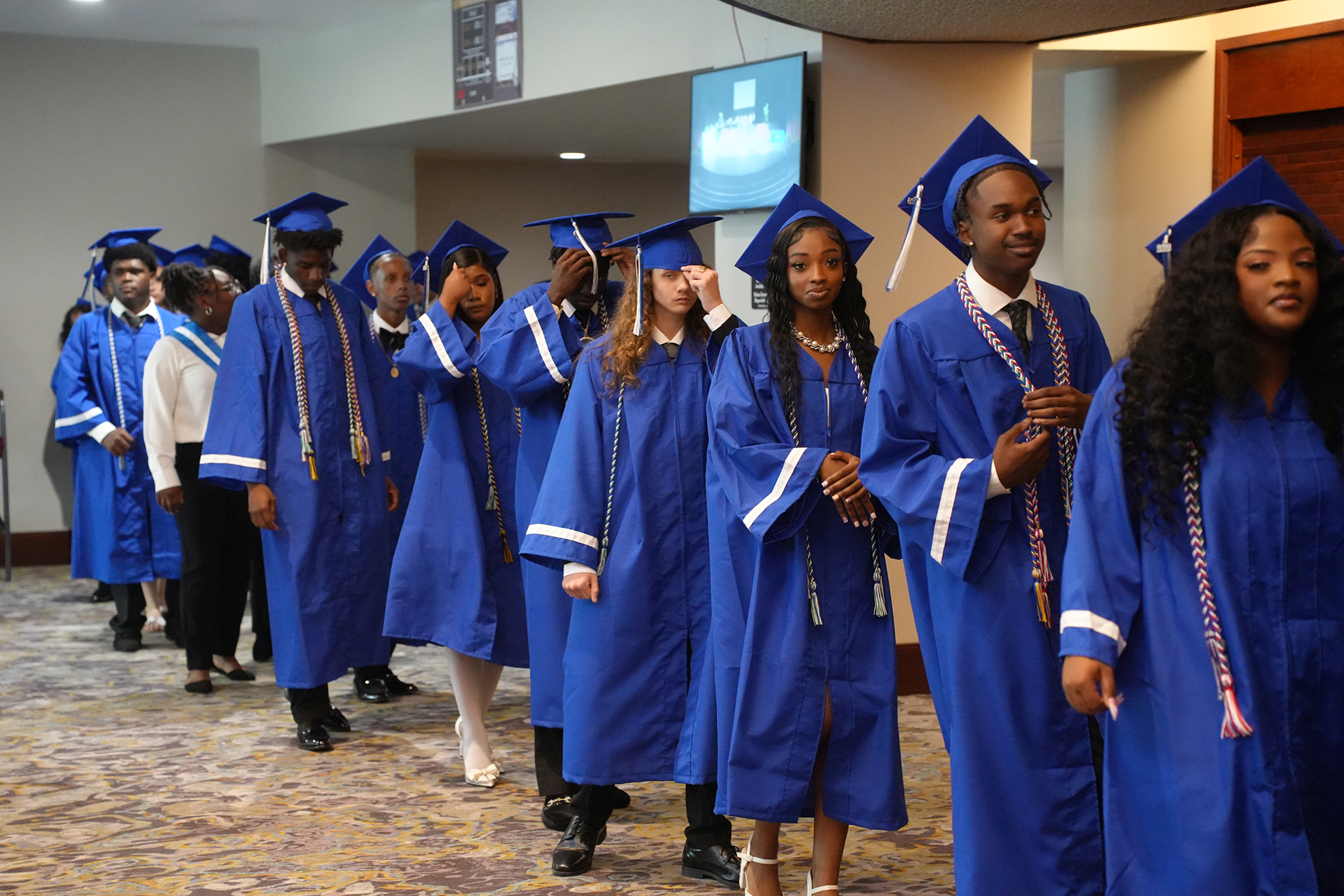 Graduates in blue gowns and caps line up in a hallway with carpeted flooring, a lit monitor on a wall.