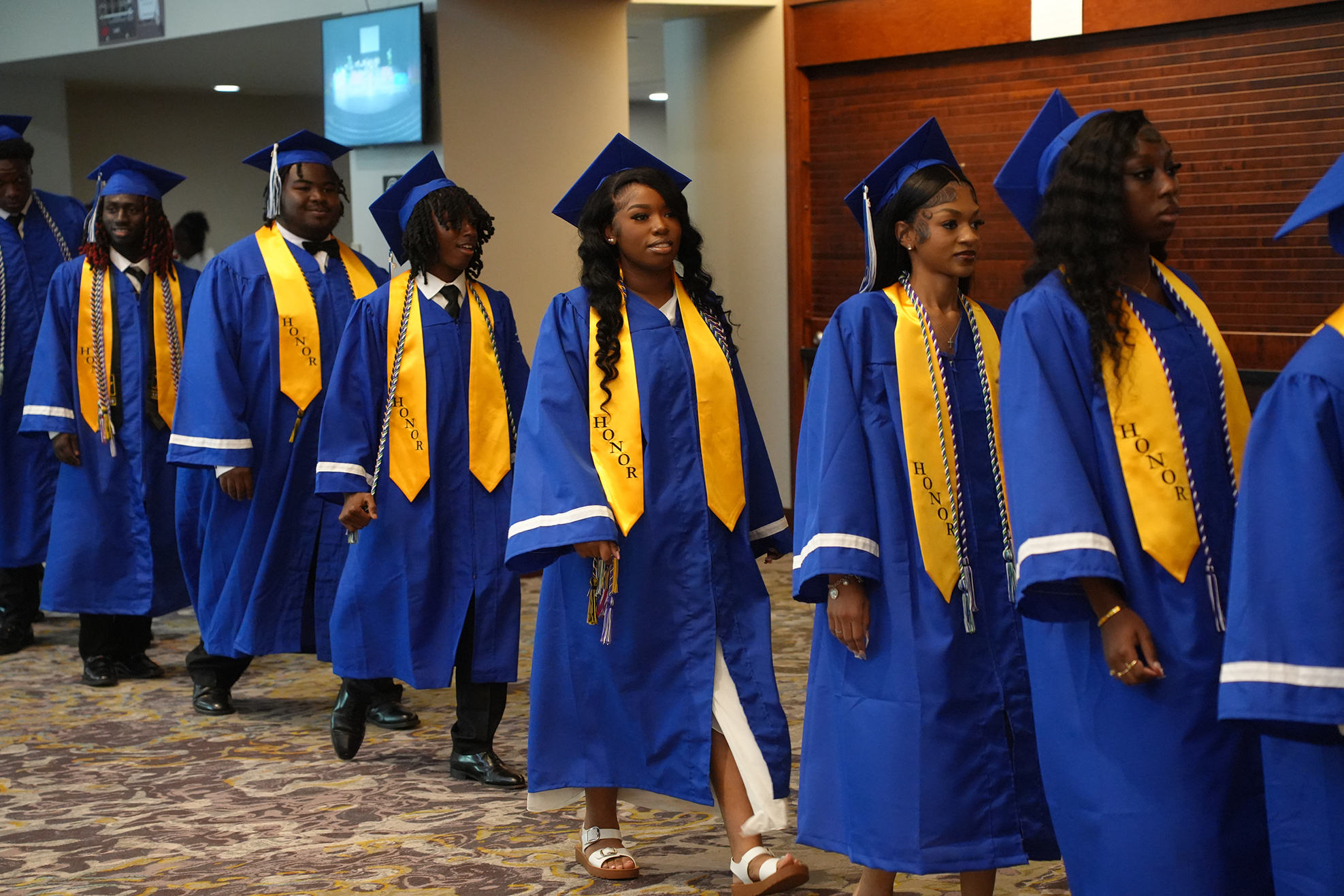 Graduates in blue and yellow gowns walk in a hallway with wood panels, tiled floors, and a screen.