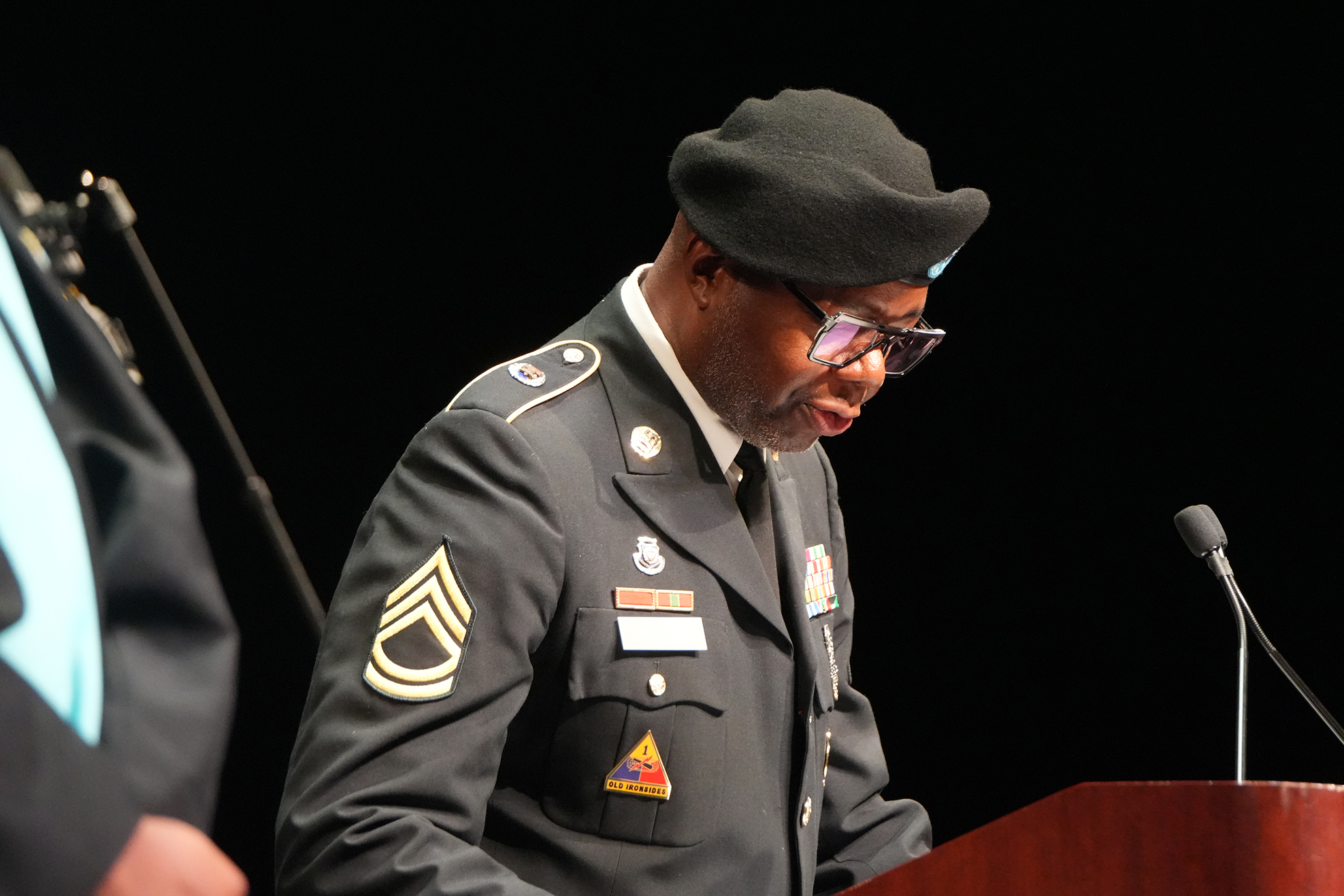 Man in military uniform, wearing a black beret, reading from a podium with a microphone.