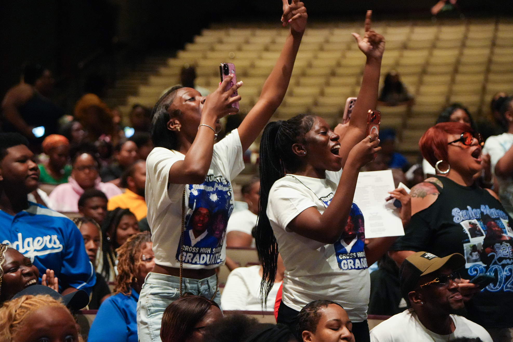 Two women in white shirts, holding phones, raise their hands and sing in a large auditorium.