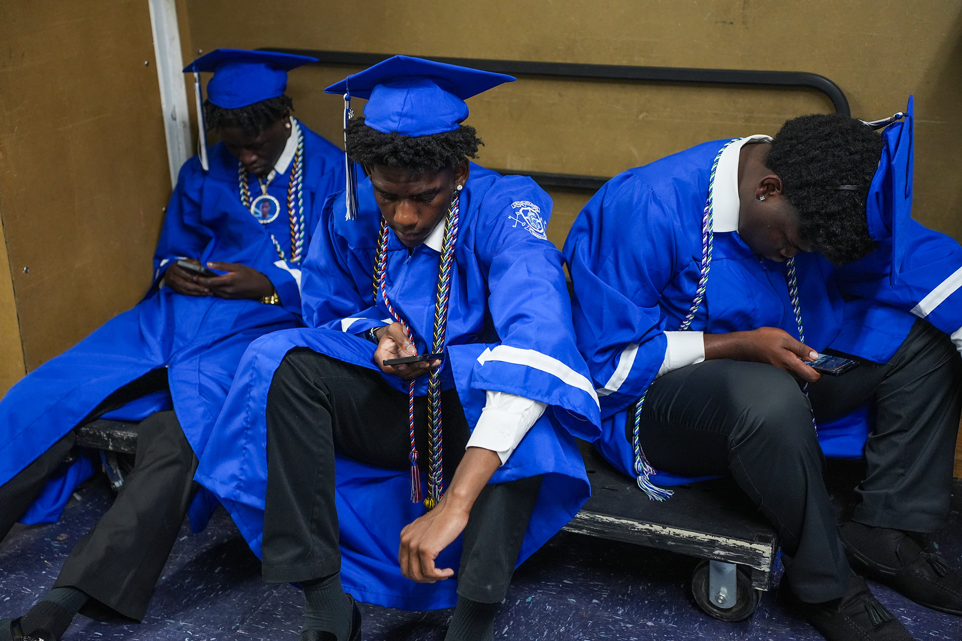 Three men in blue caps and gowns sit on a bench, each looking at a smartphone.
