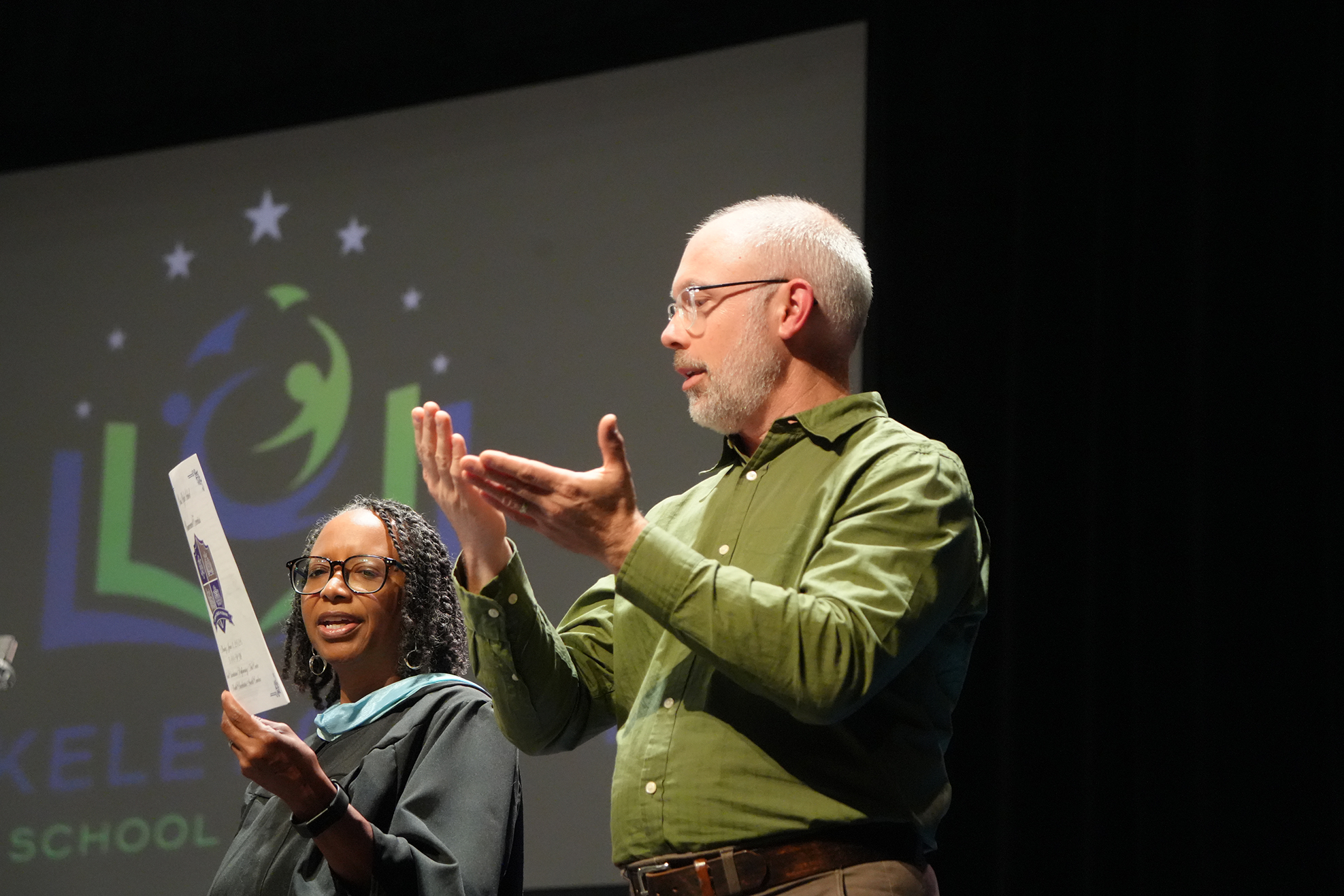 Man and woman facing each other. Man speaks with raised hands. Woman holds a paper. Behind them, a projector screen displays a logo.