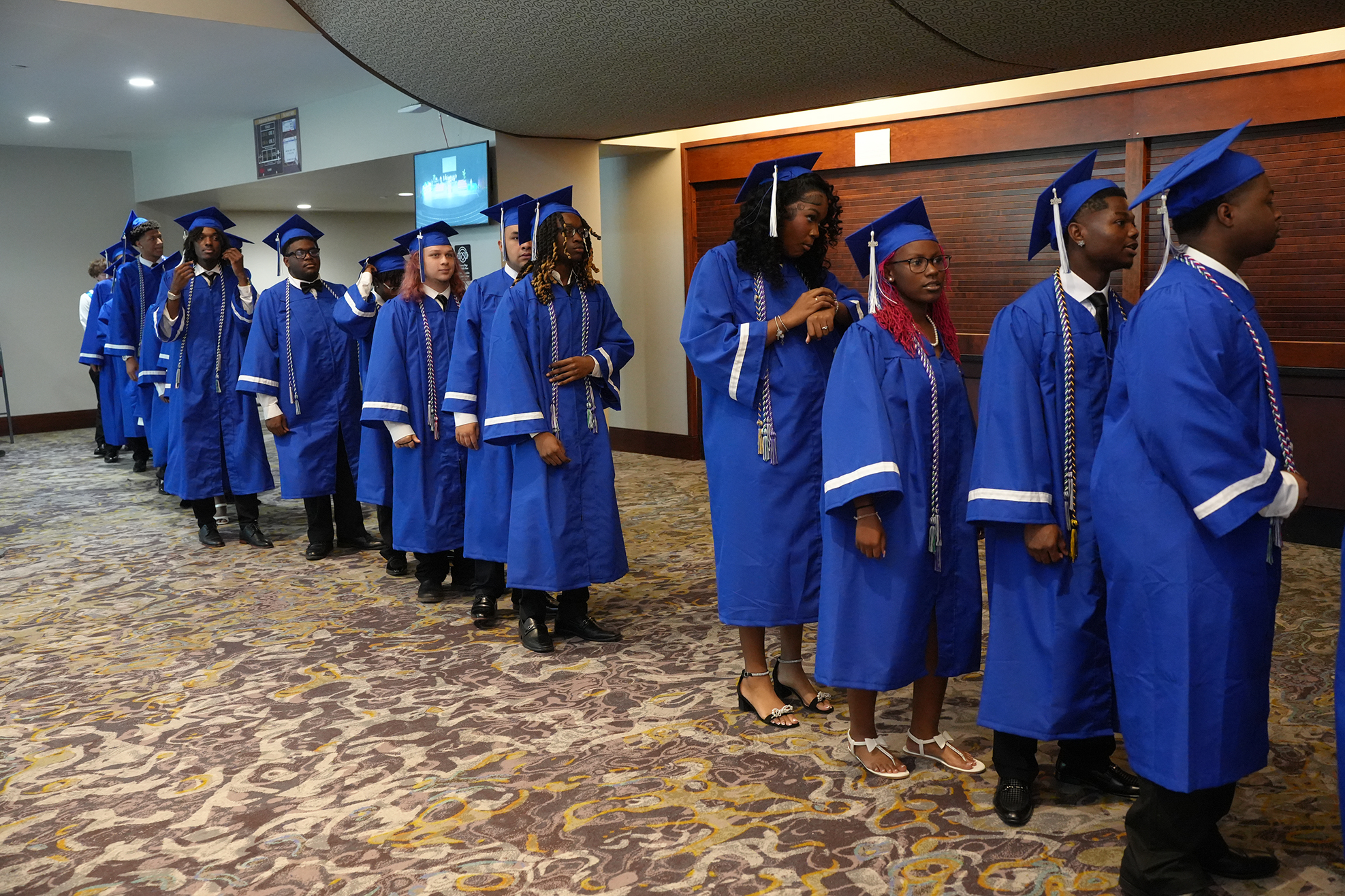 A row of students in blue graduation gowns and caps standing in a hall, facing forward.