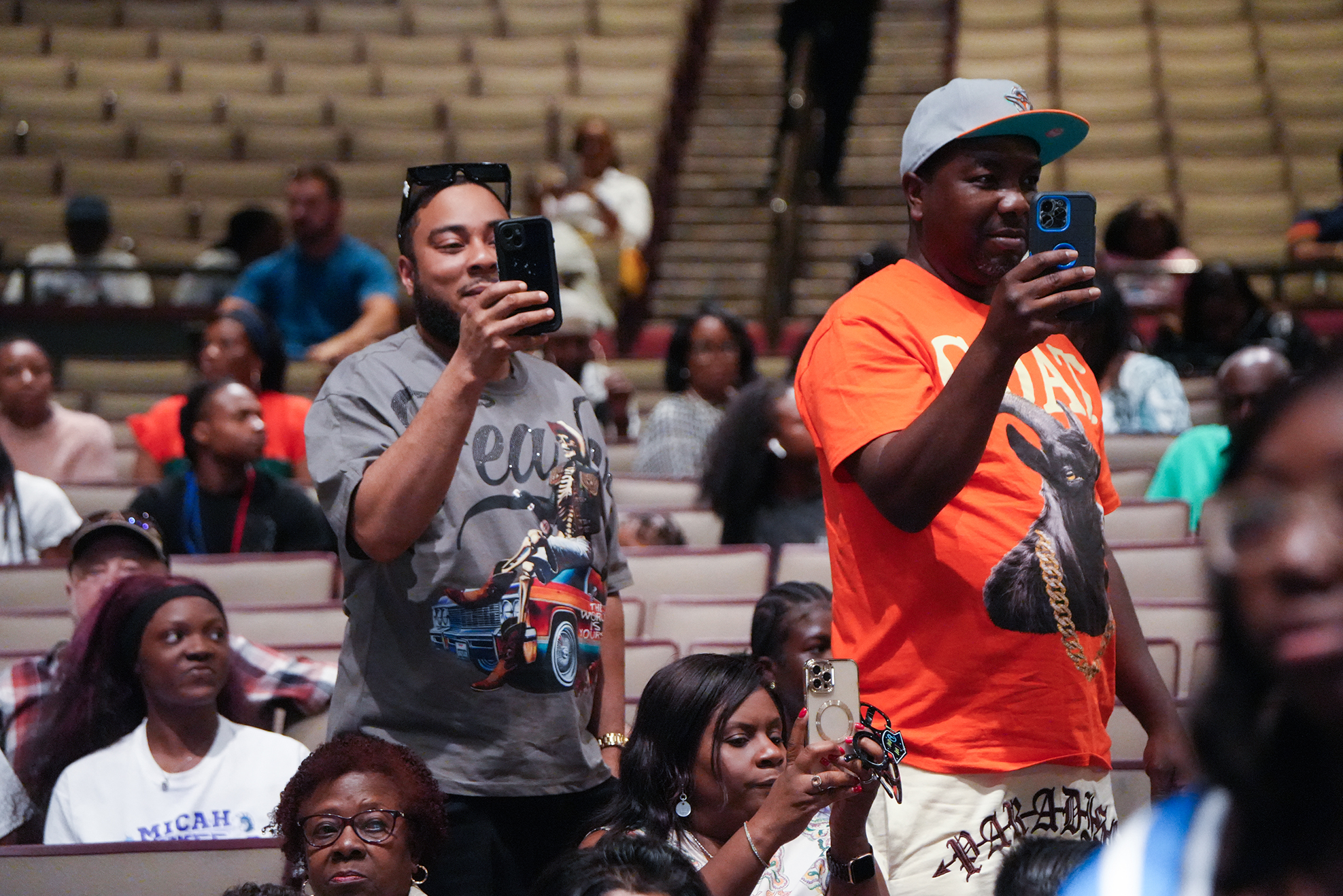 Audience members in a theater, two men in front using cellphones to take photos, with blurred people in the background.