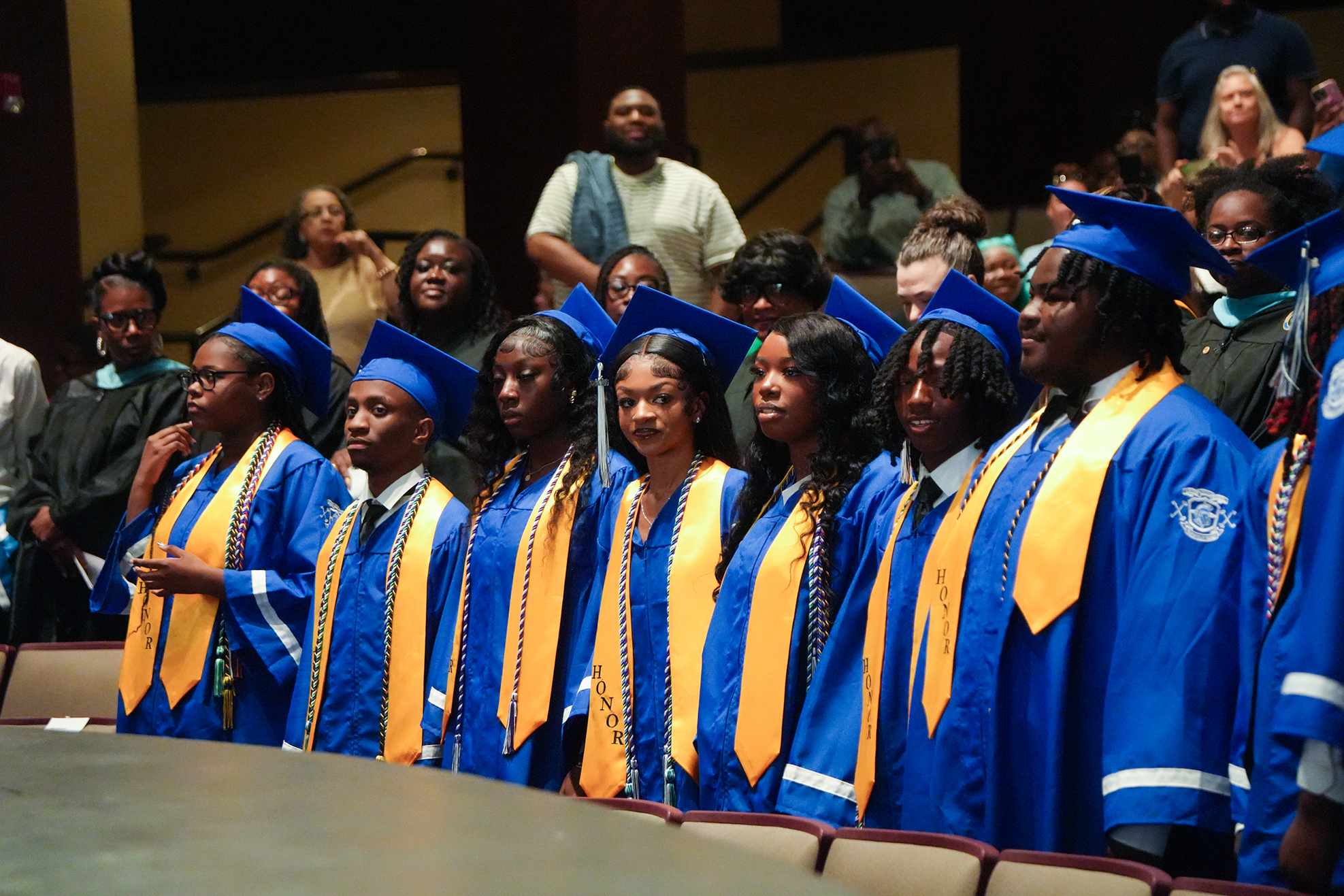A row of students in blue and yellow graduation gowns and caps standing on stage in a theater.