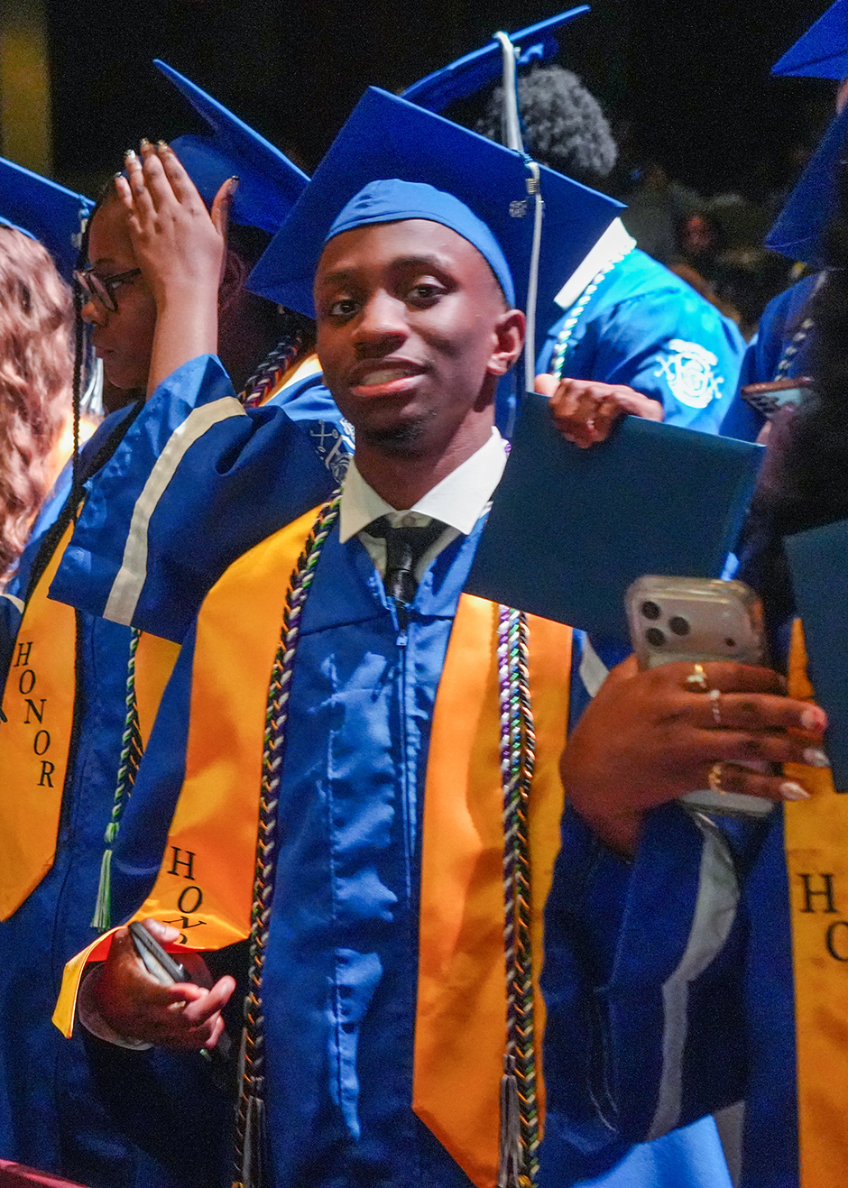 People in blue and gold graduation gowns with tassels and hats stand together. One holds a folder and a cellphone.