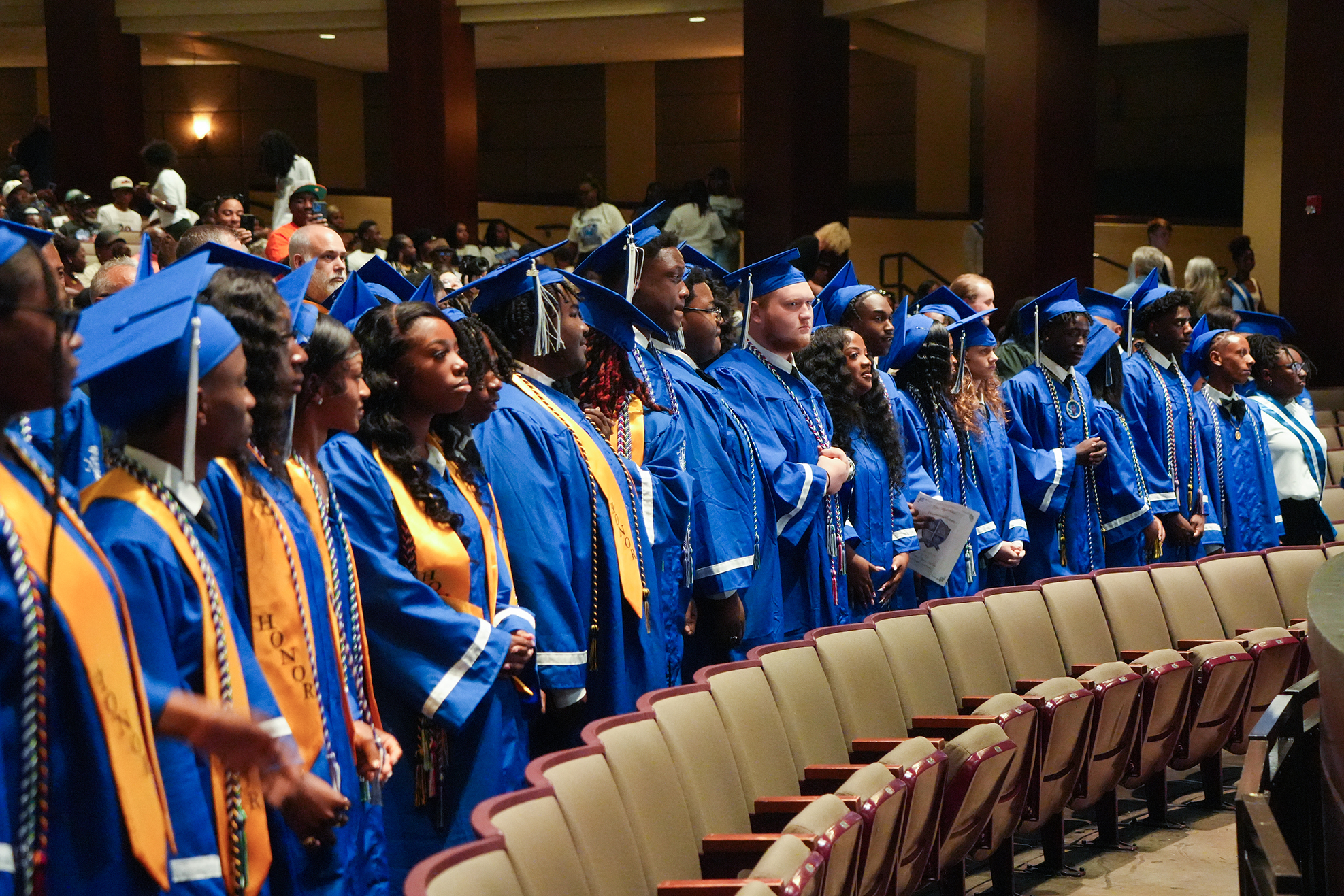 A row of people in blue and gold graduation robes stand in an auditorium, facing forward.