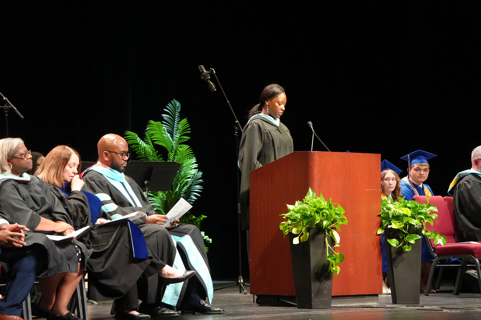 People in caps and gowns sit in chairs on a stage. One woman stands at a podium.