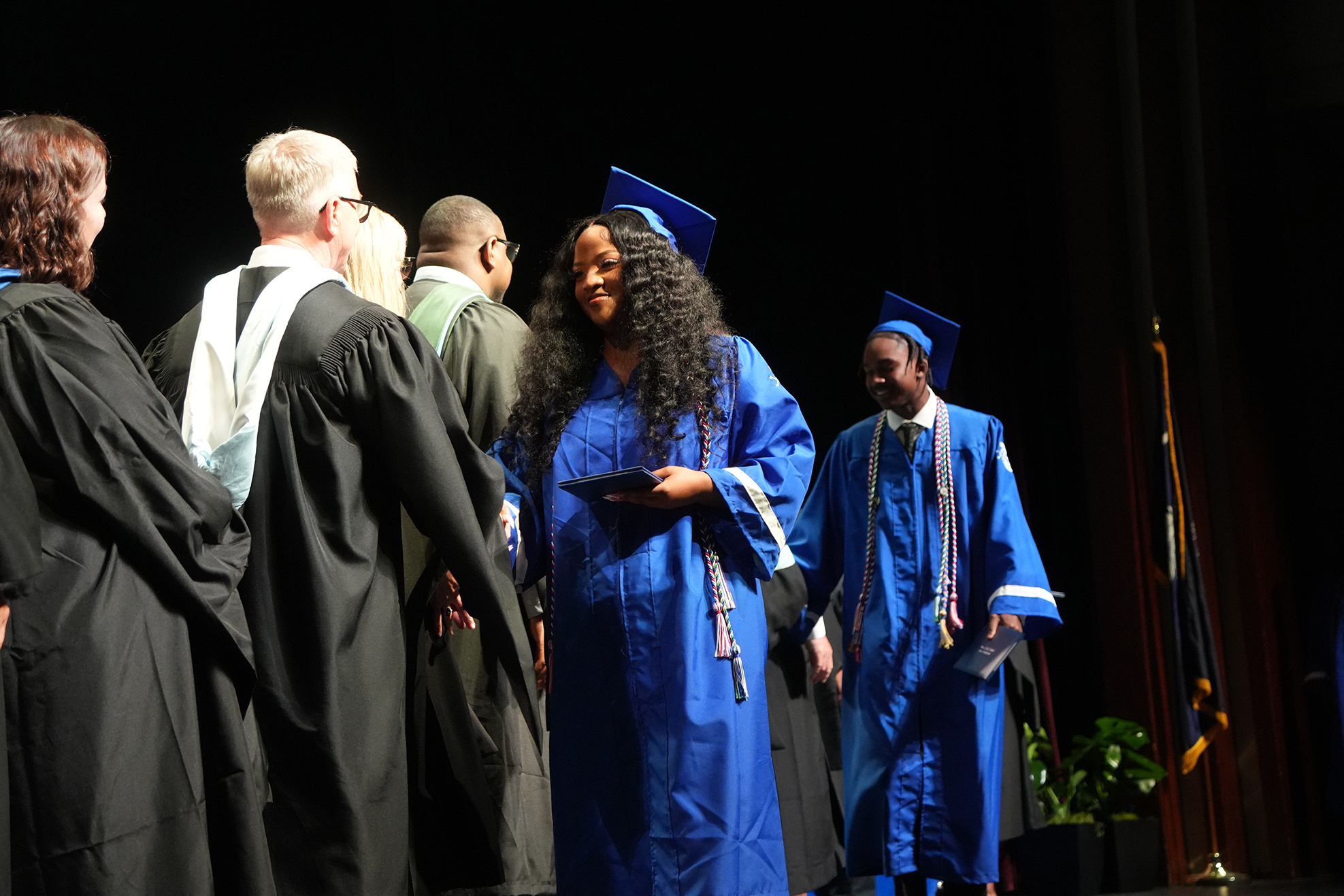 A stage with people in blue and black gowns. A woman in a blue gown with a tasseled hat holds a diploma.
