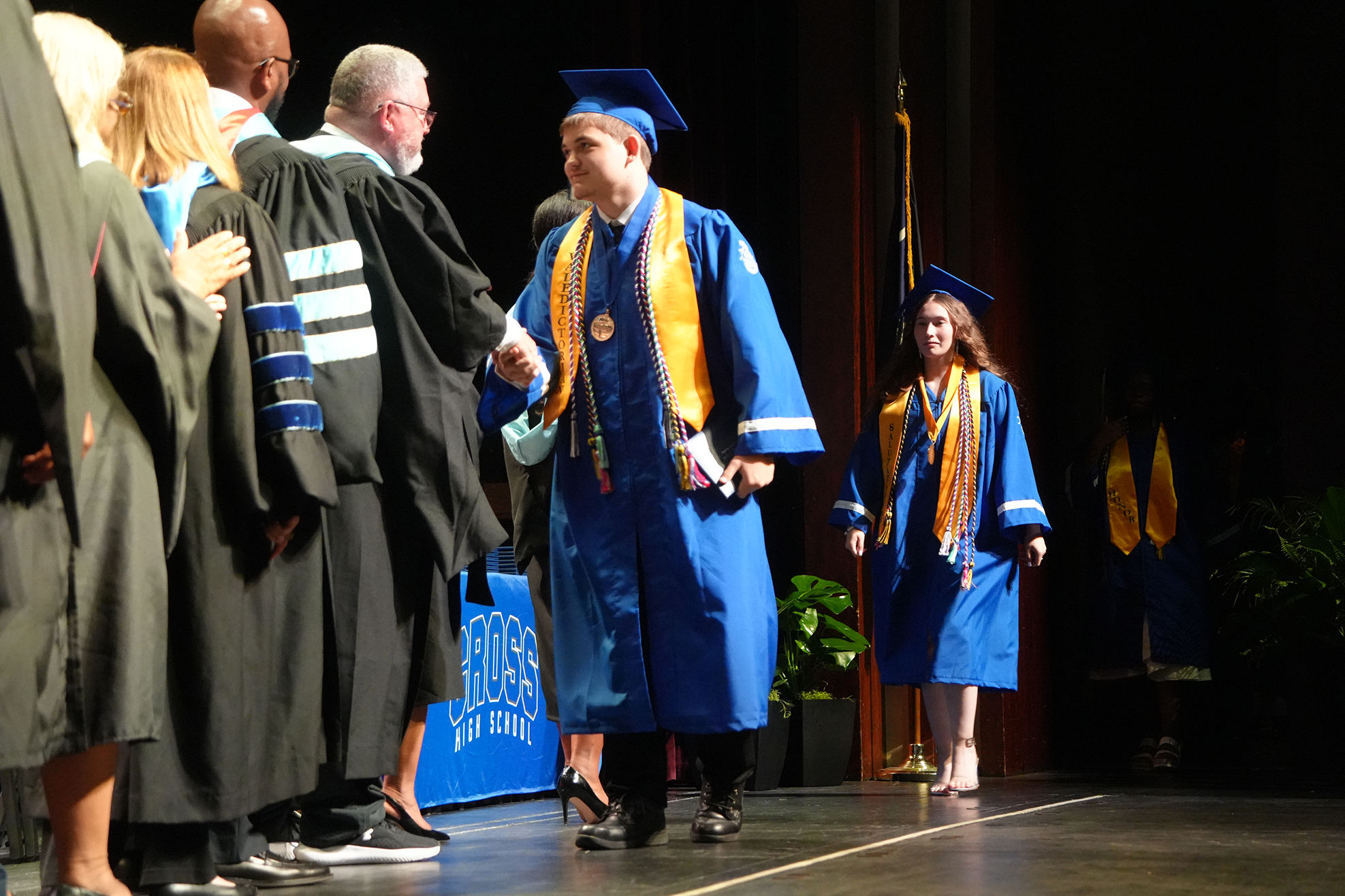 A group of people on a stage, one man in a blue gown, another in a black gown. A woman in a blue gown approaches.