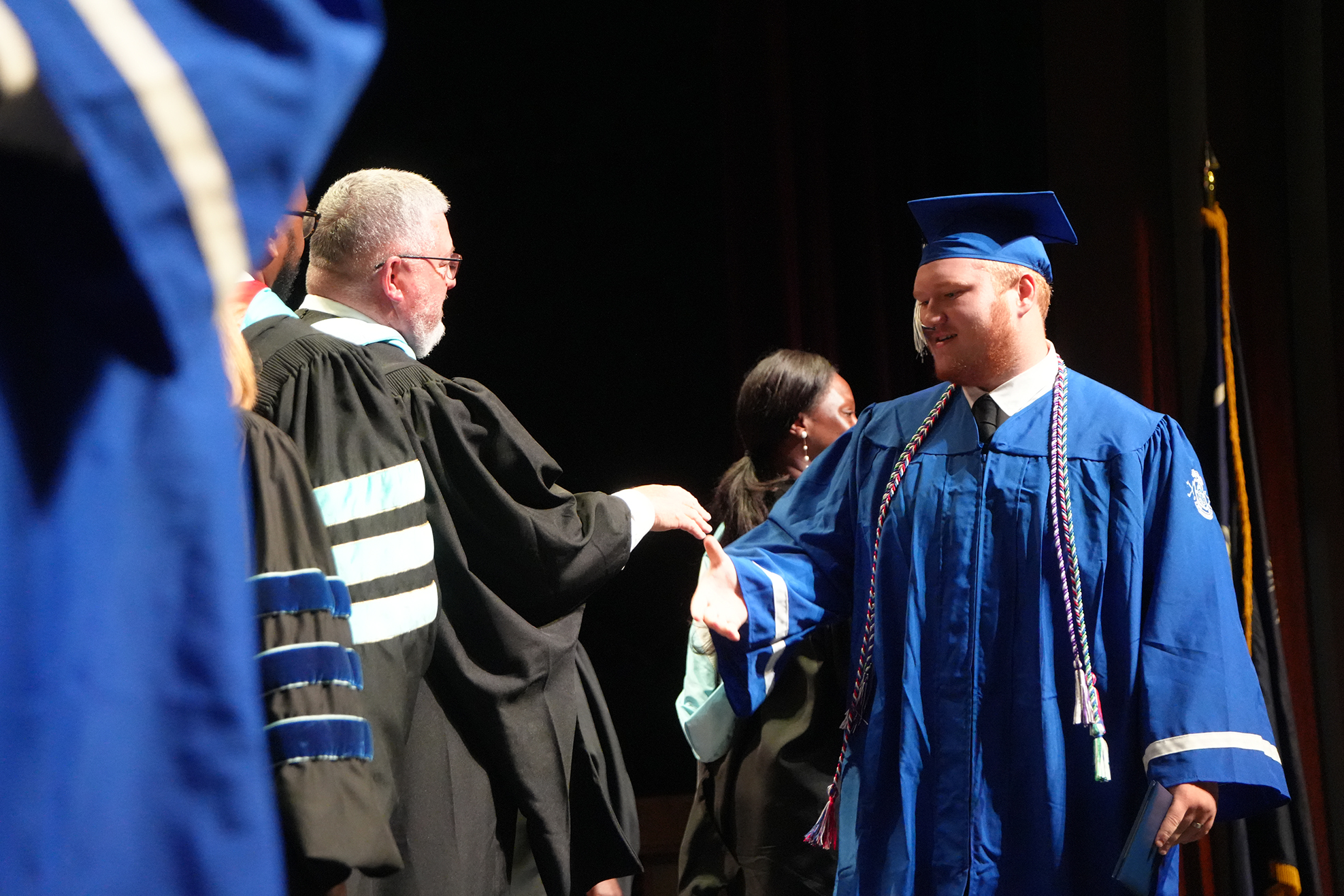 A person in a blue graduation gown shakes hands with a faculty member on a stage.
