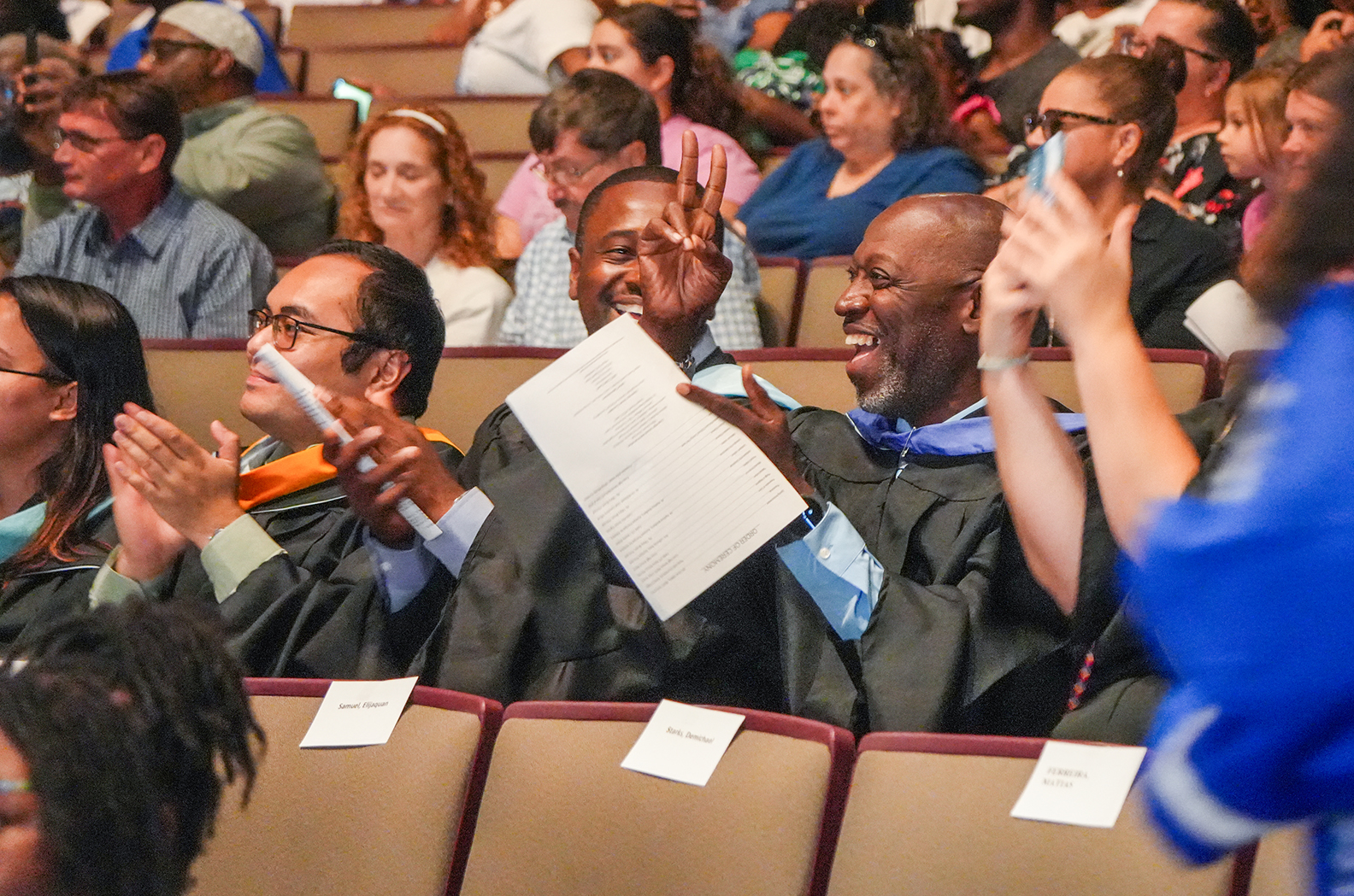 A group of people in academic attire, smiling and clapping, in a theater-like setting with rows of seats.