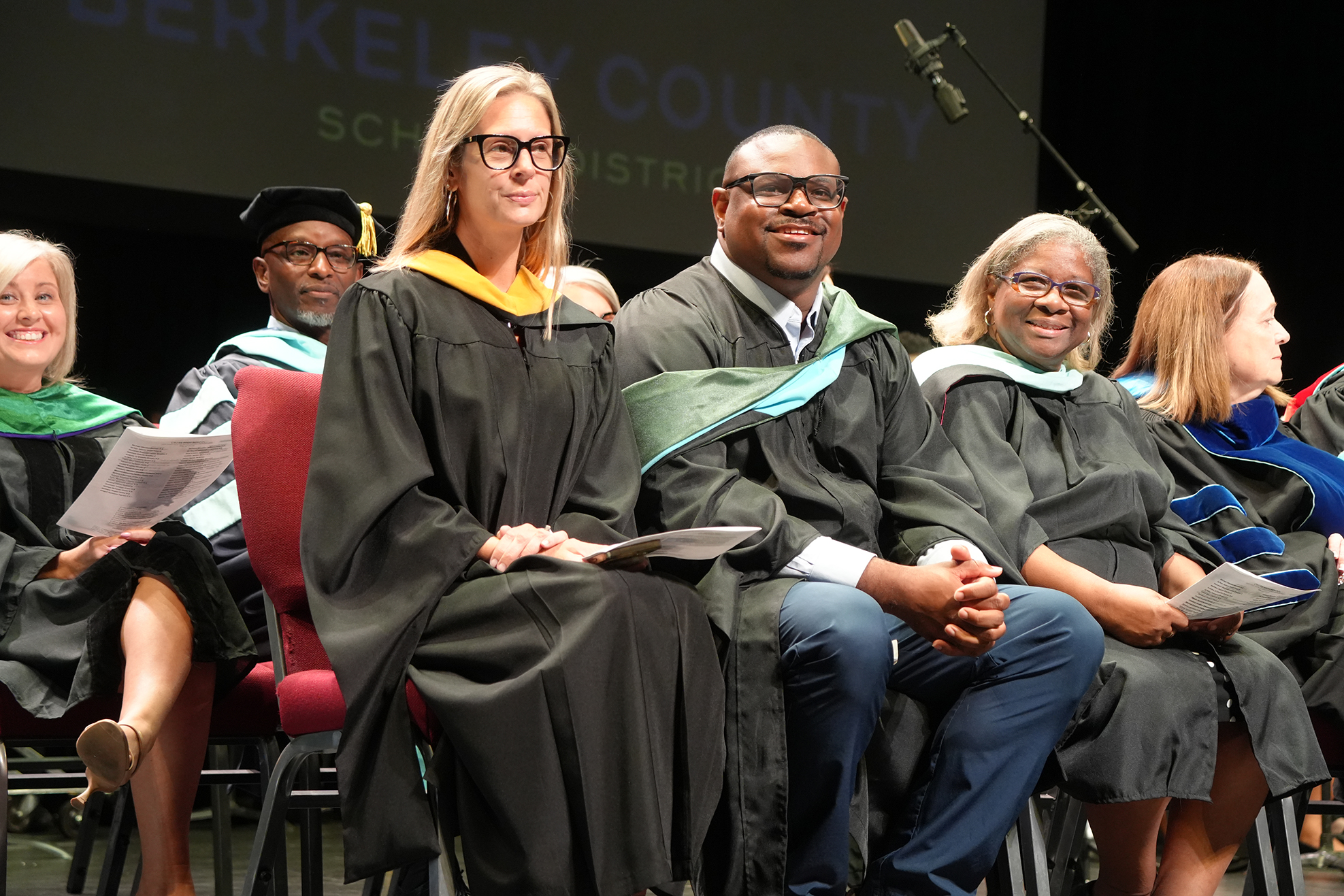 Graduates in academic regalia sit on a stage, possibly during a ceremony. A large screen behind them reads "Berkeley County."