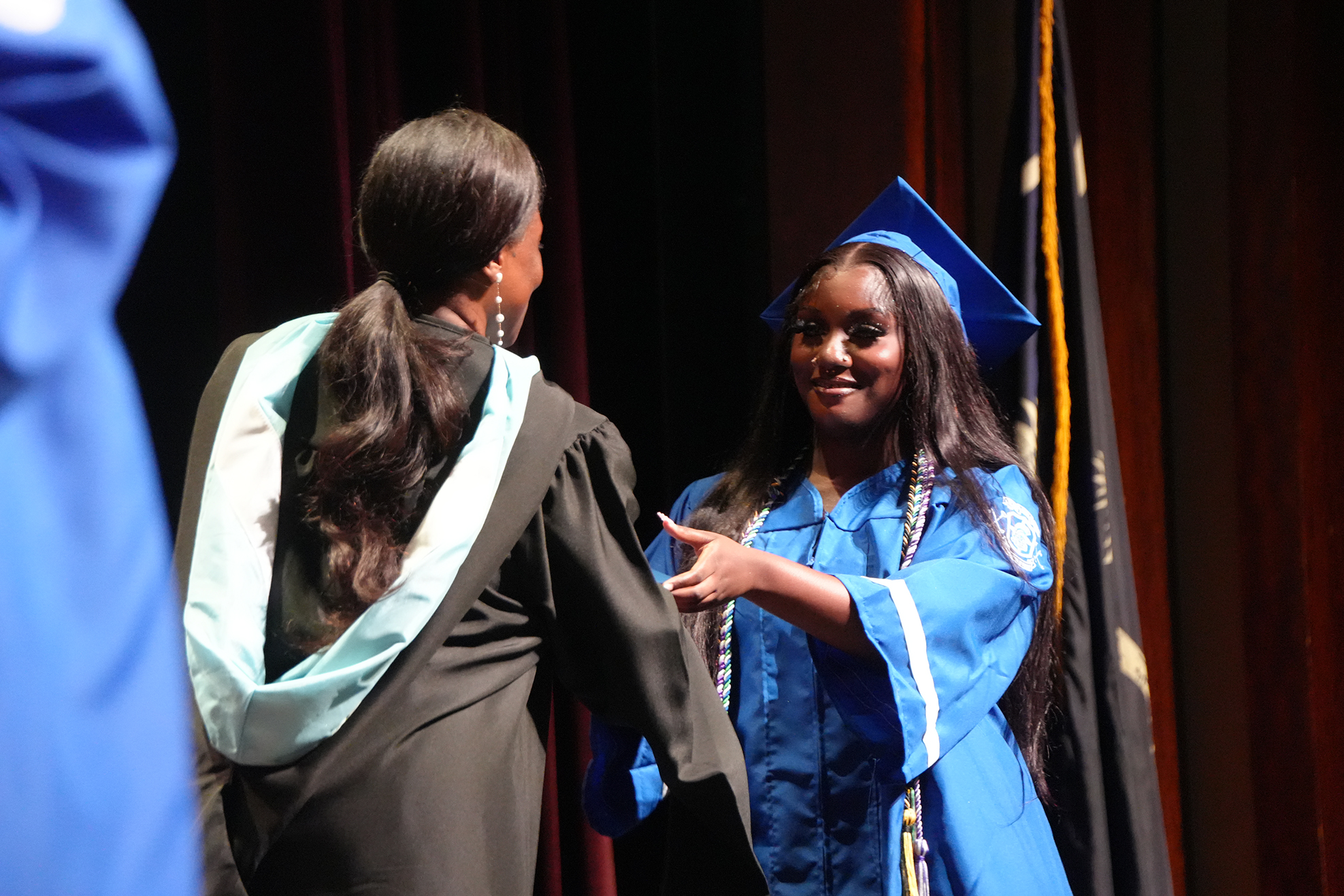 Two women in academic robes, one wearing a cap and gown, converse on stage with a flag in the background.