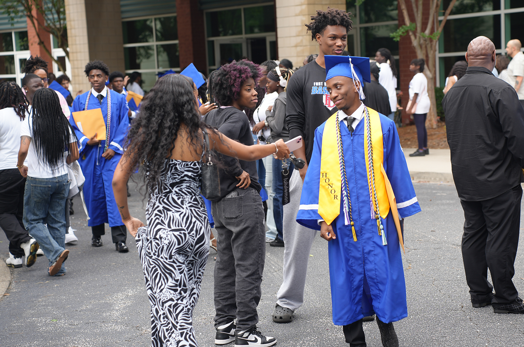 People in graduation attire gather outside a building; a man in a blue gown shakes hands with a woman.