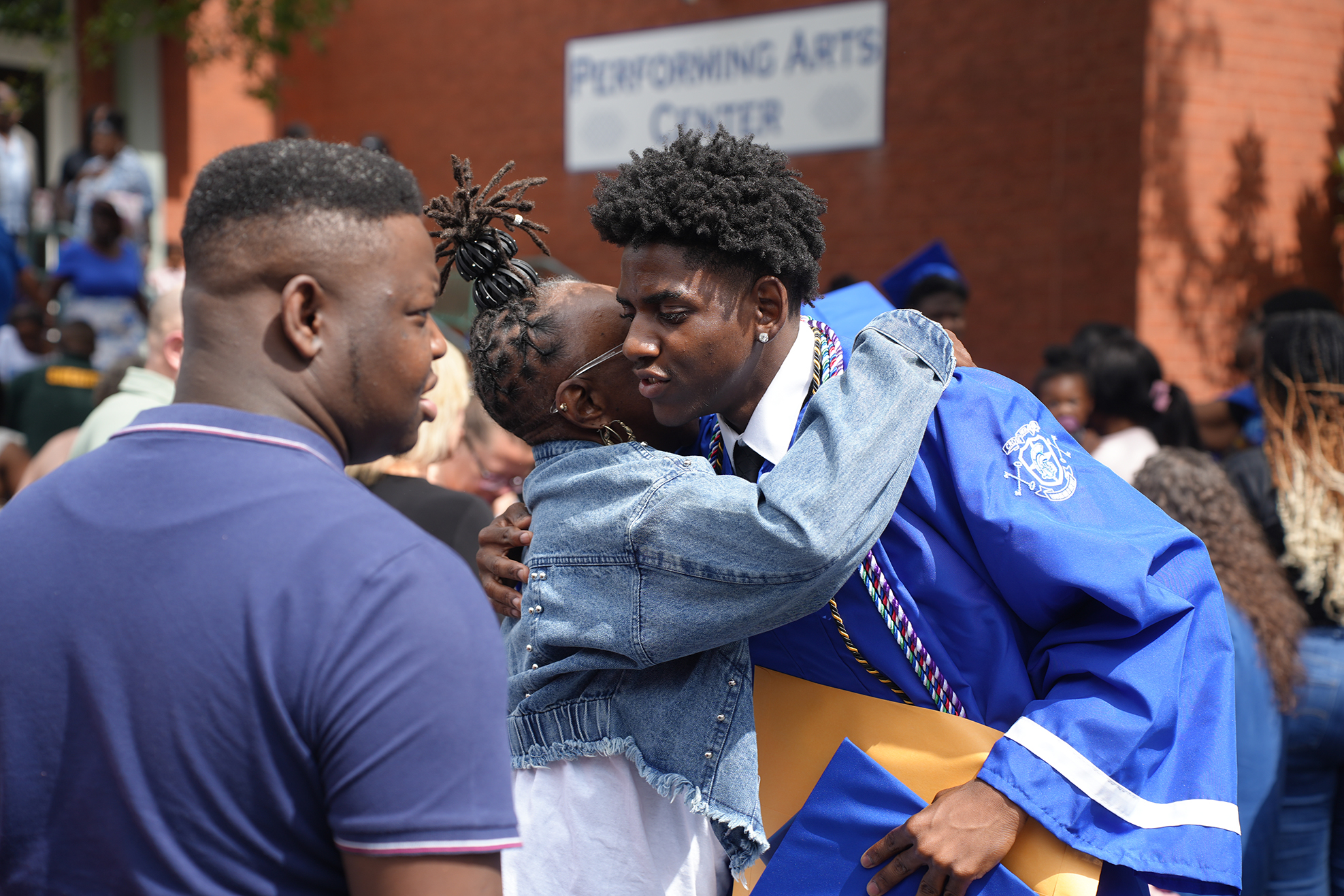 Two adults and a child embrace outside. A young man in a blue graduation robe hugs a child.