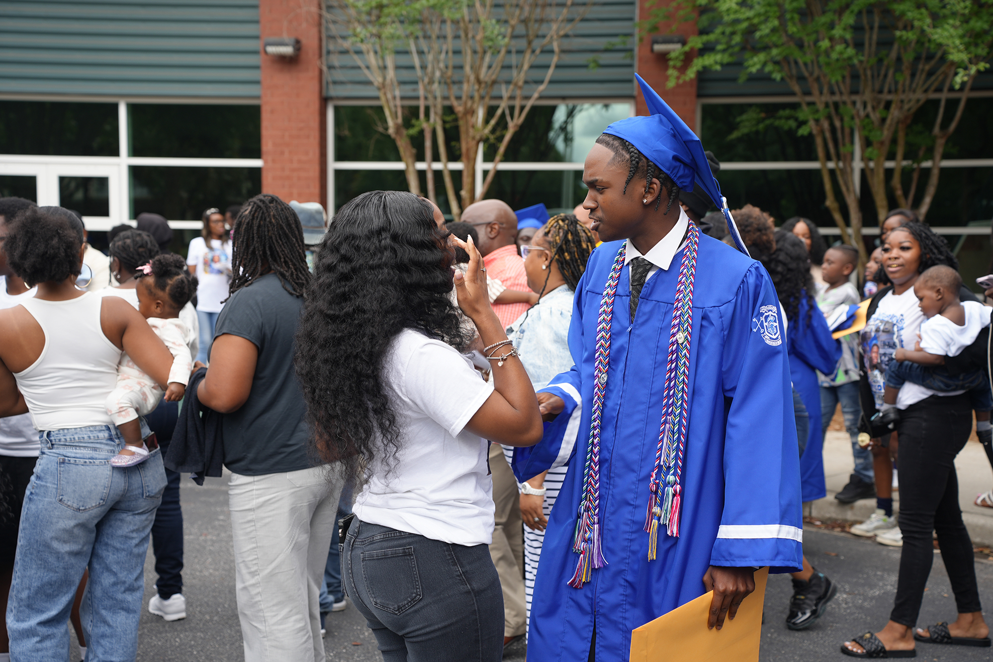 A group gathers outdoors; a man in a blue graduation gown holds a folder and converses with a woman.