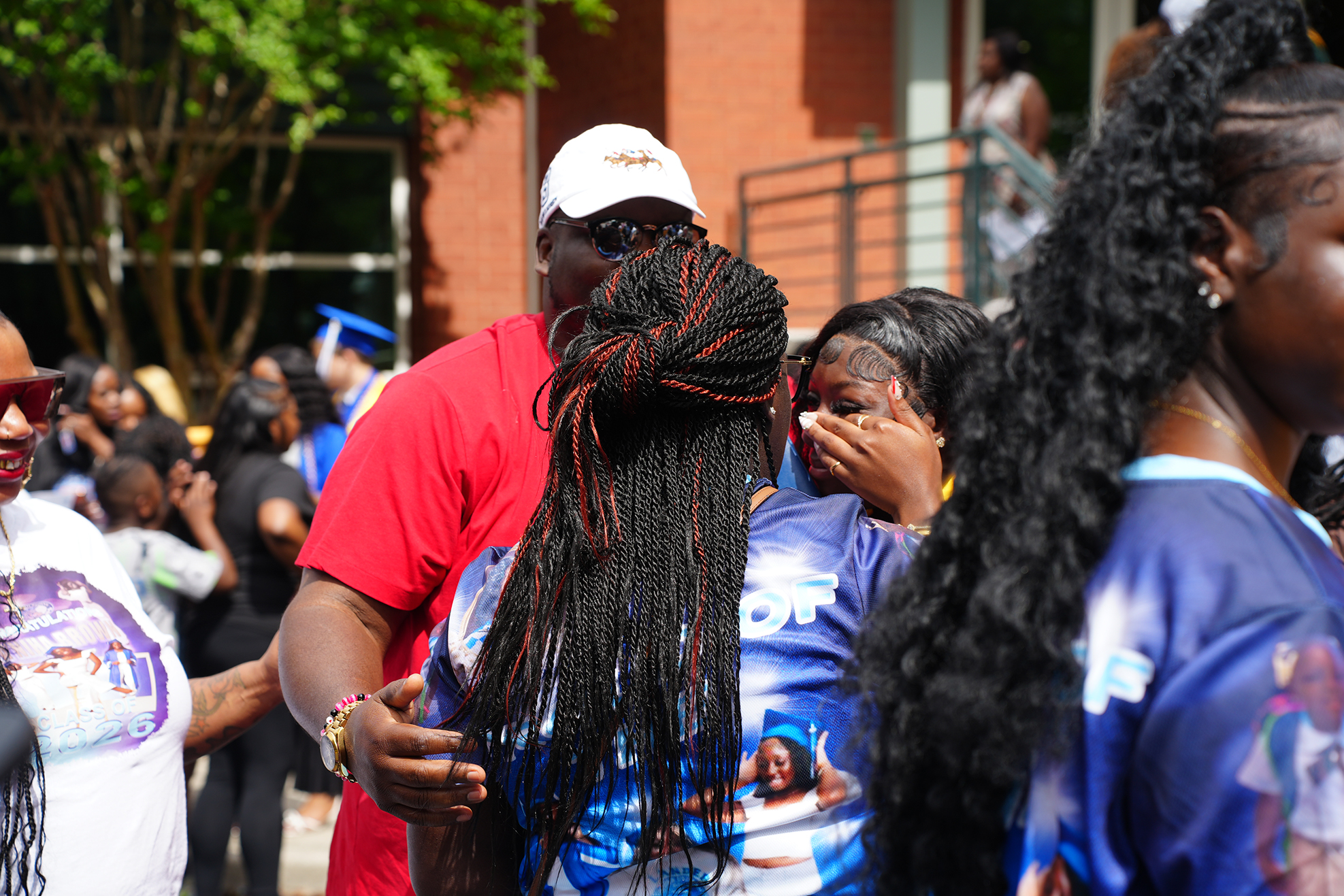 People gather outside a building. A man in a red shirt supports a woman with long braids.