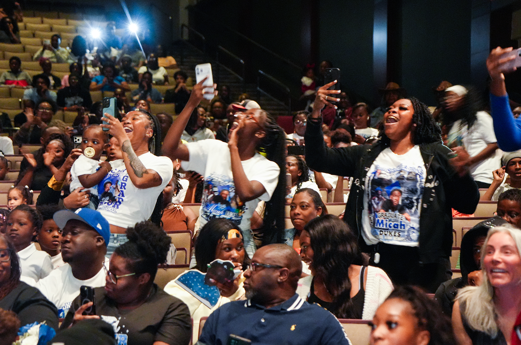 A crowd of people in an auditorium, some wearing matching white shirts, raising hands and holding phones to record.