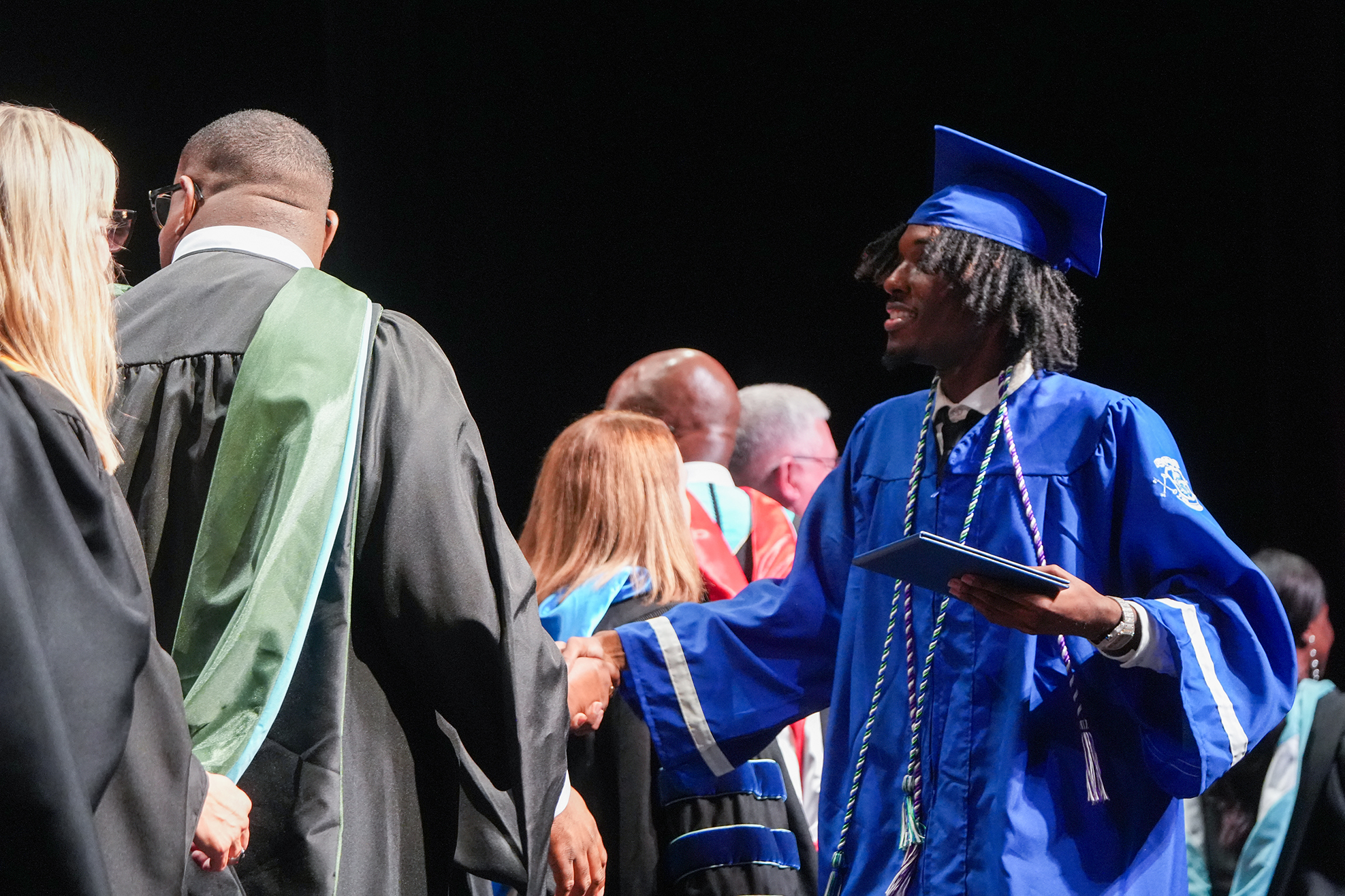 A man in a blue graduation gown and cap holds a diploma in front of several people, including a man in a green and black gown.