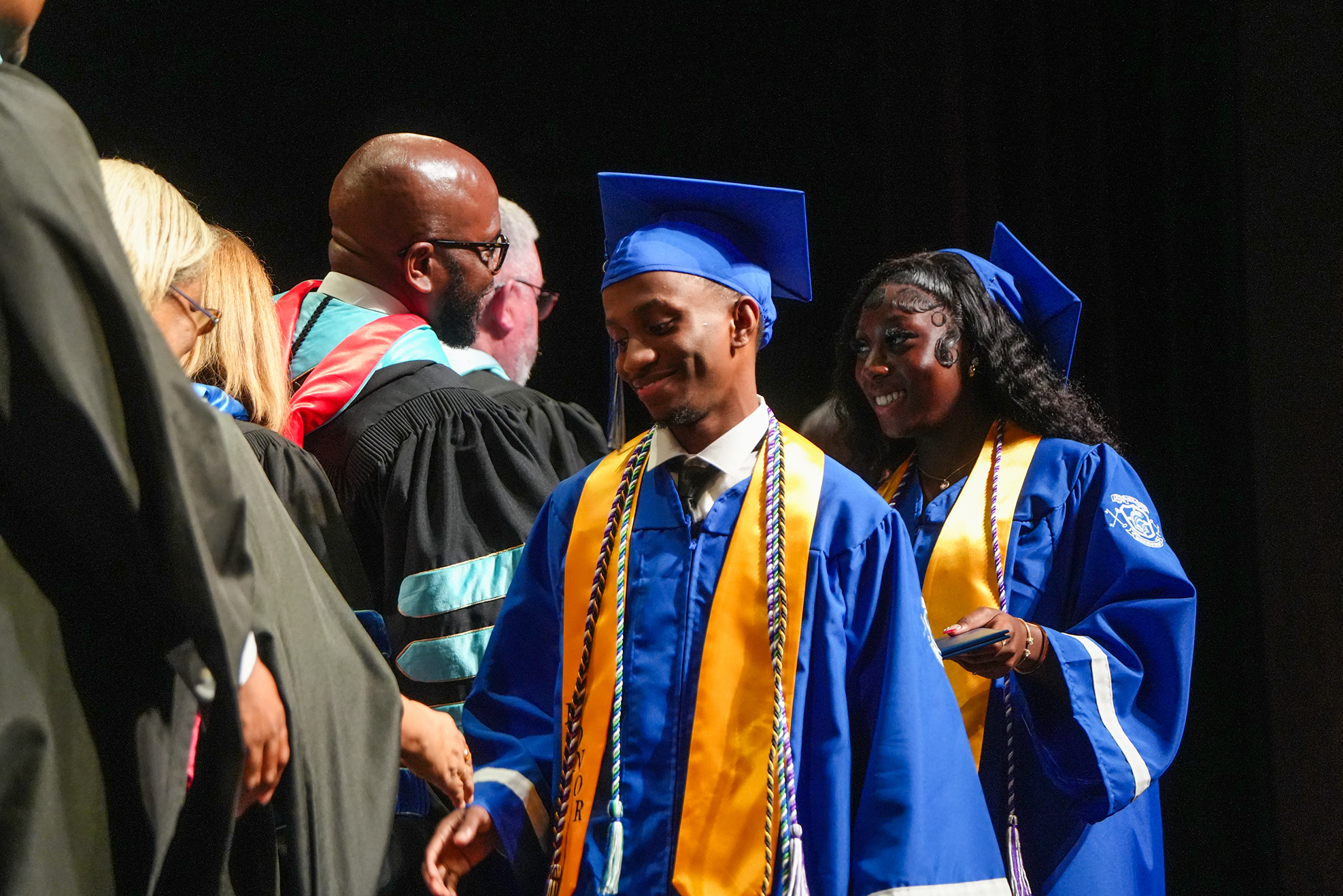 A man in a blue and yellow graduation gown and cap walks past others on stage.