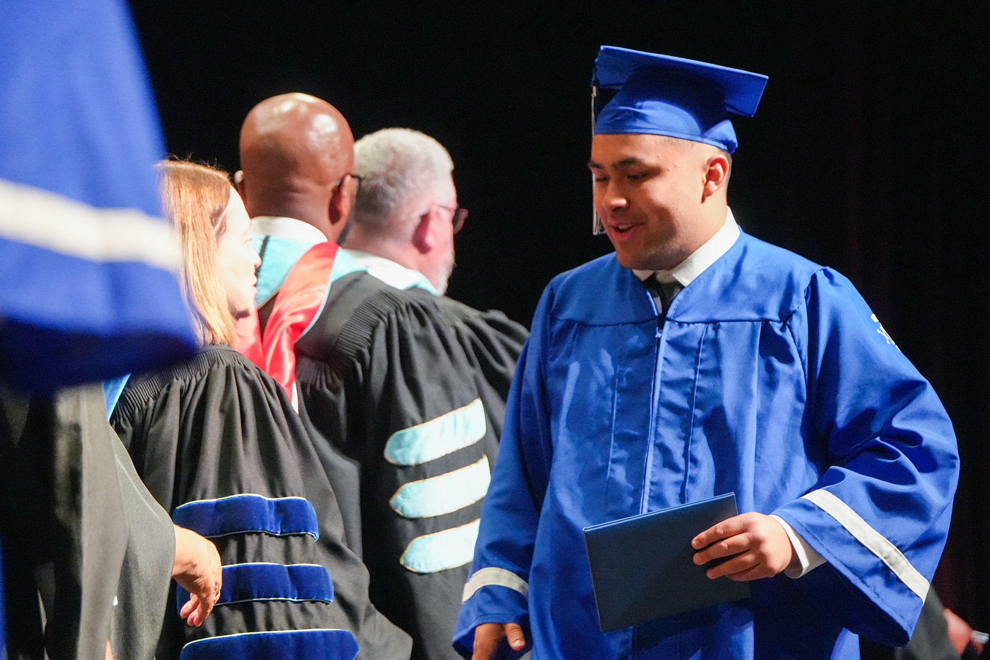 A graduate in a blue gown stands with a folder in a room with other people wearing caps and gowns.