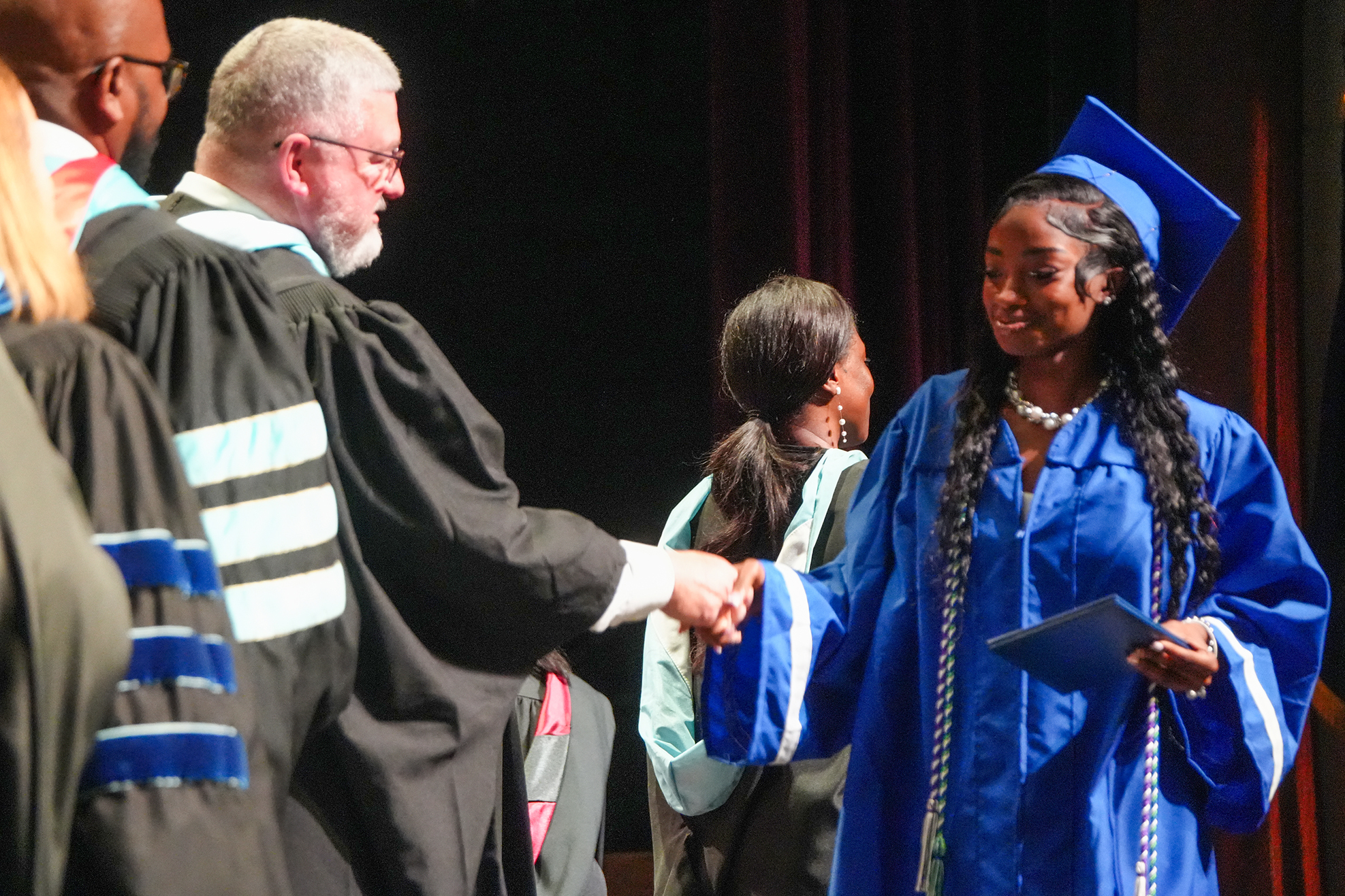 Graduates at a ceremony, man in academic gown shakes woman's hand. Woman wears blue gown, holds diploma.