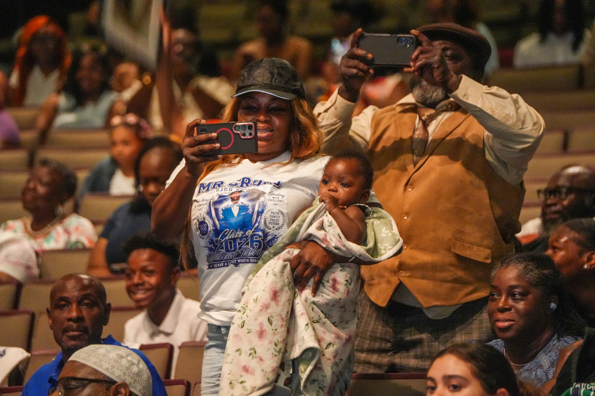 A group of people seated in an auditorium; a woman in a cap holds a baby, while a man behind her holds a phone to record.