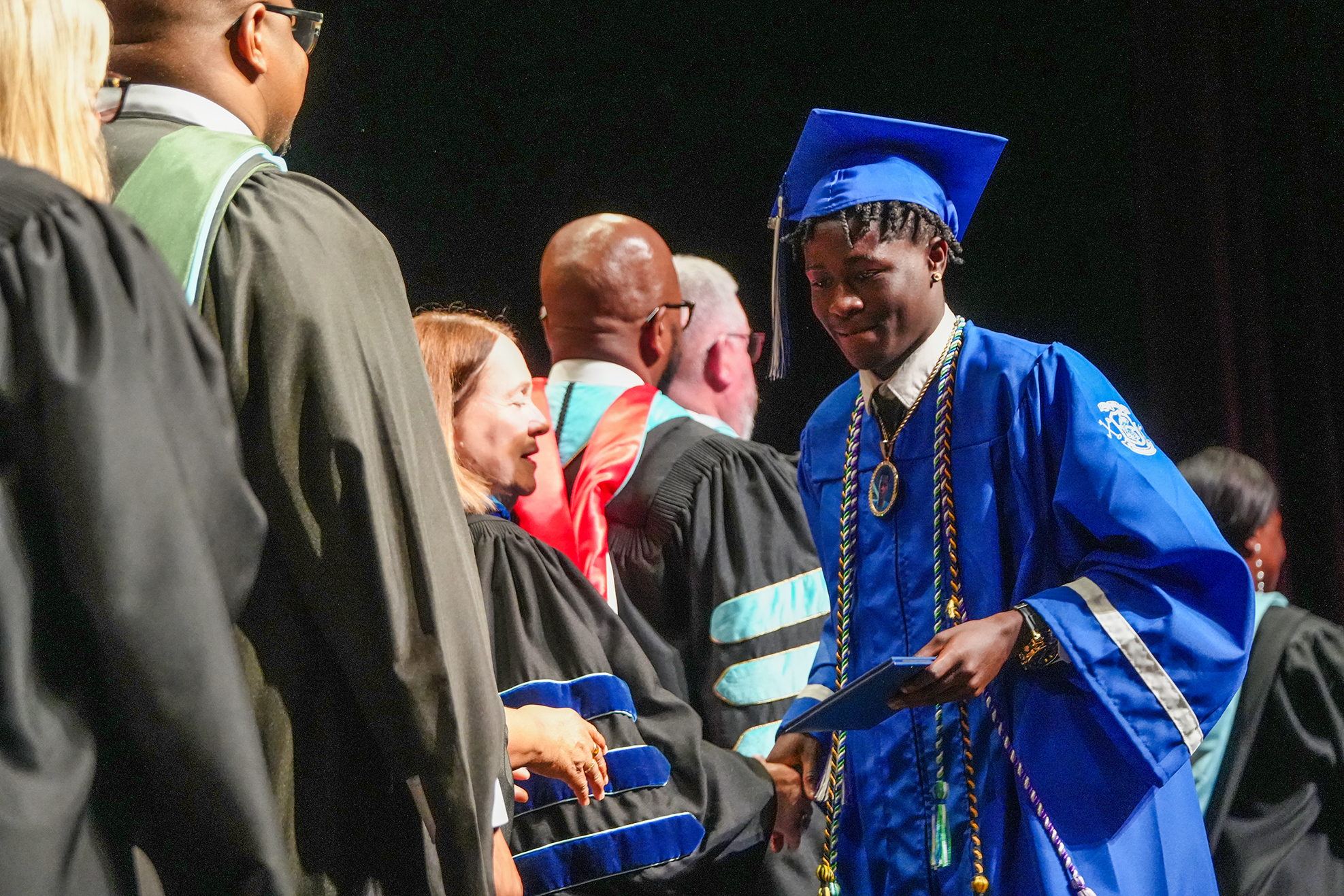 Several individuals in academic regalia stand together. A man in a blue gown and cap looks at a paper.