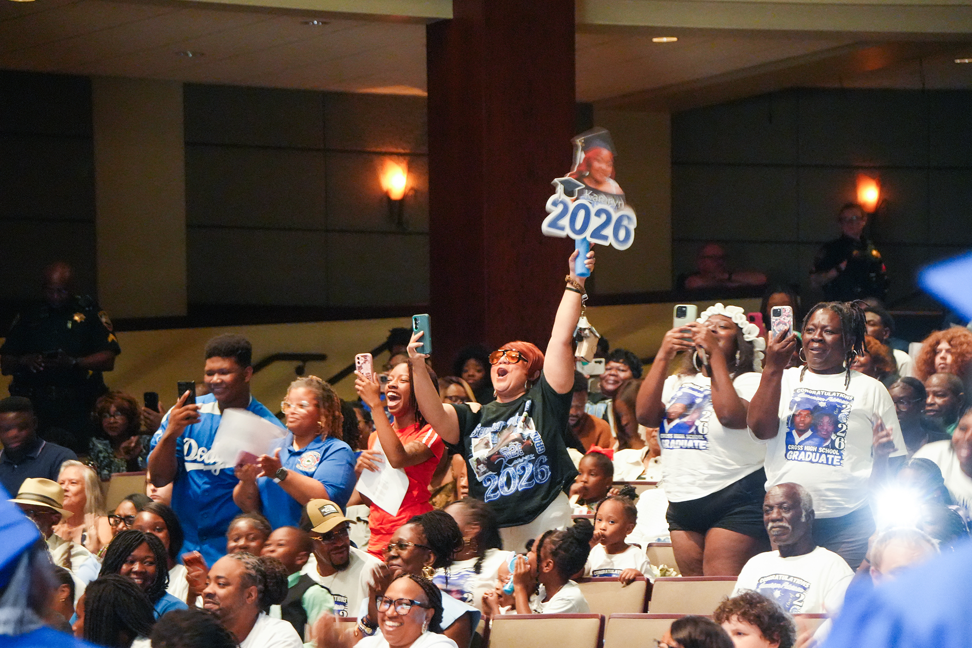 A crowd of people in a large room, many holding up phones and signs. A person holds a sign reading "2026."