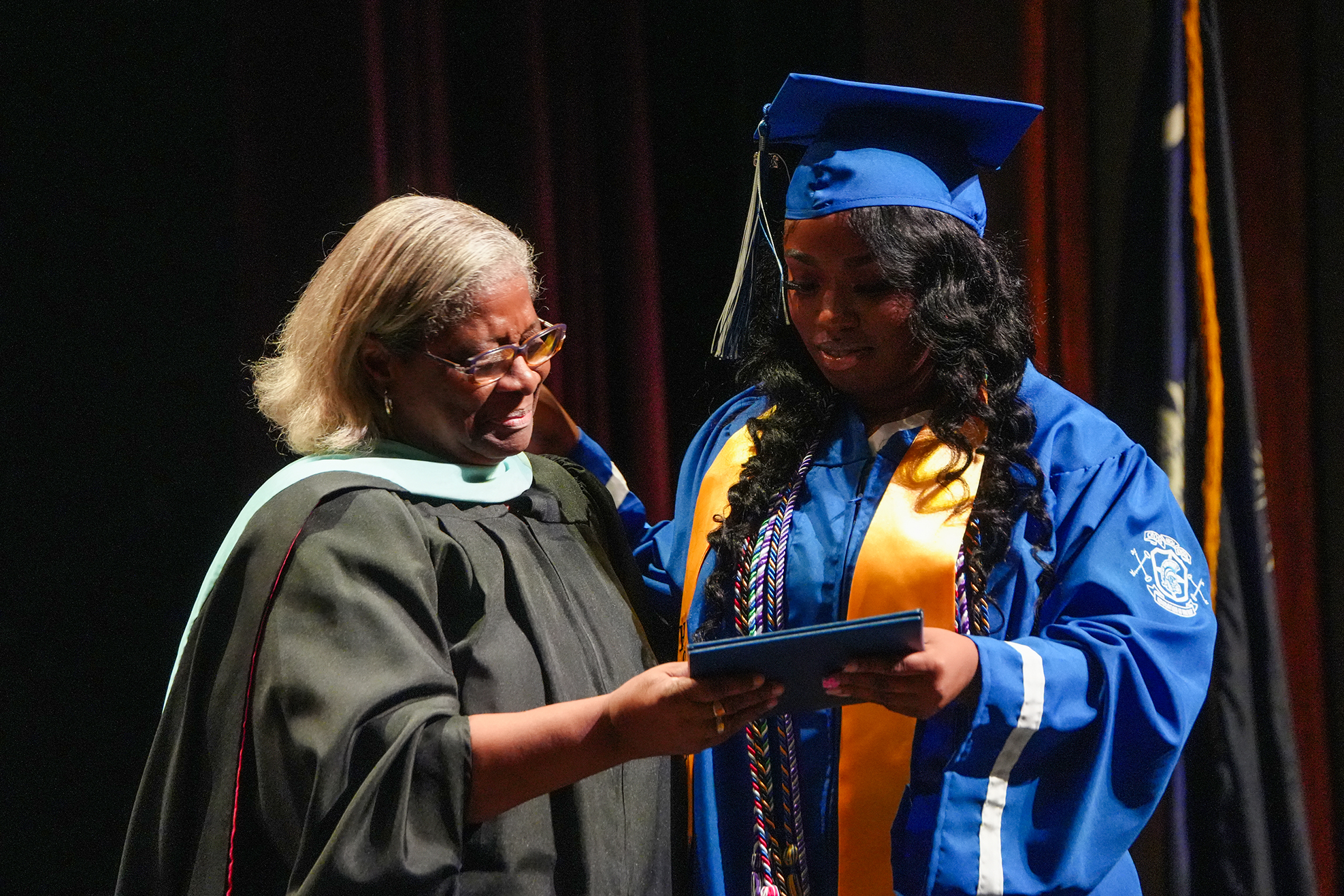 A woman in a blue gown, cap, and sash stands with a woman in a black gown and glasses, both holding a diploma.