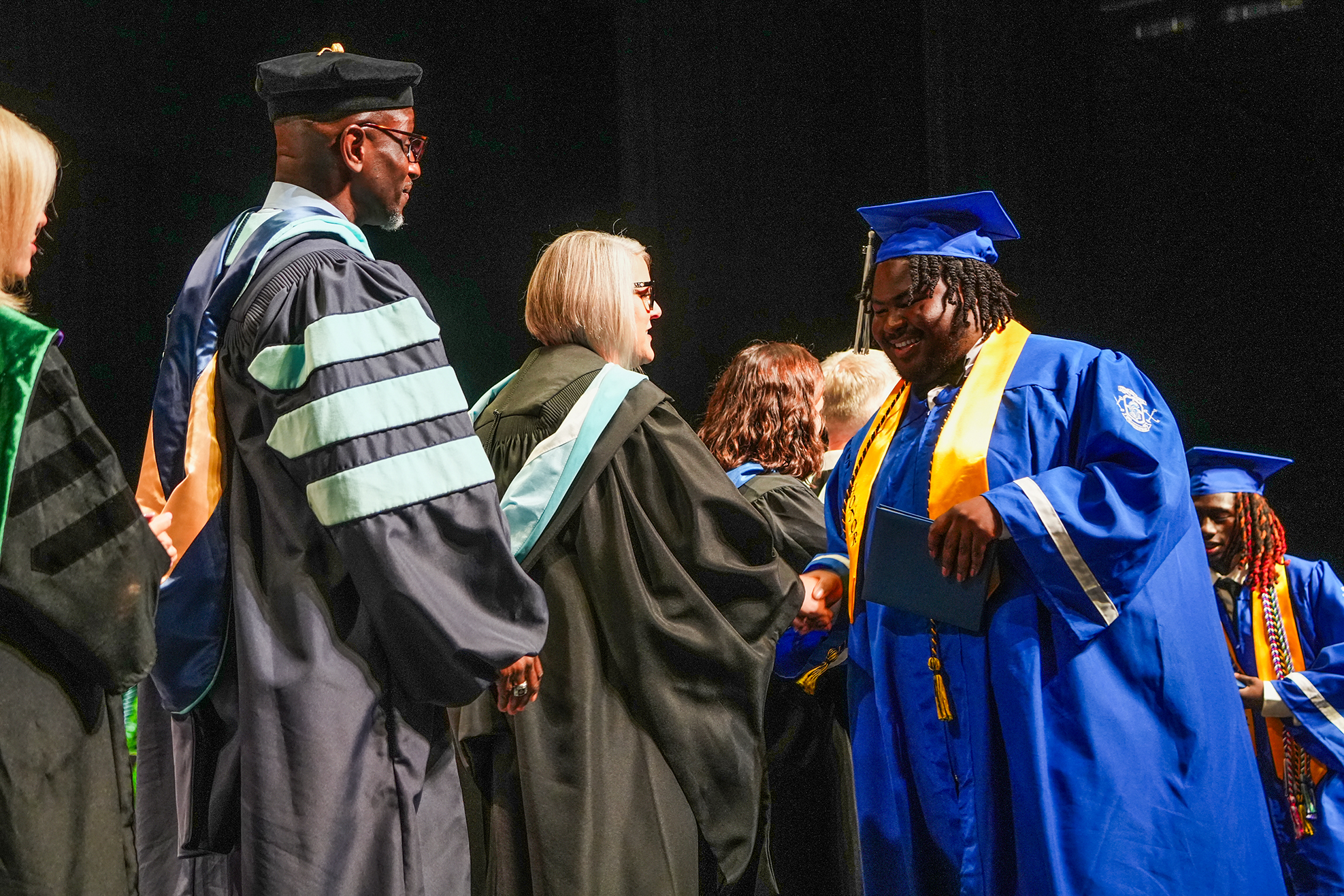 Graduates in blue and black gowns, one in blue with a gold sash, conversing with faculty at an event.
