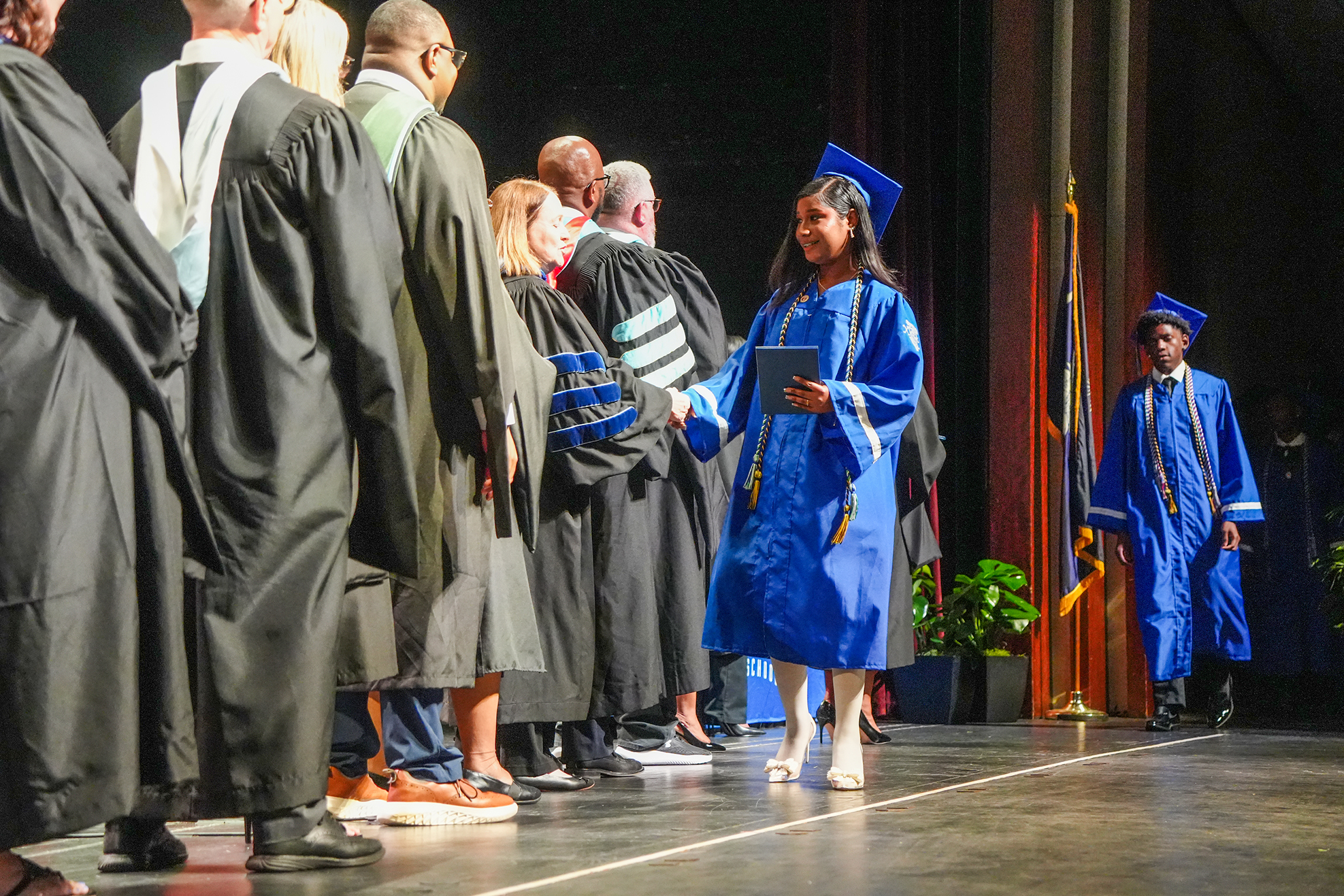 A young person in a blue gown walks toward others on stage in front of a backdrop with plants.