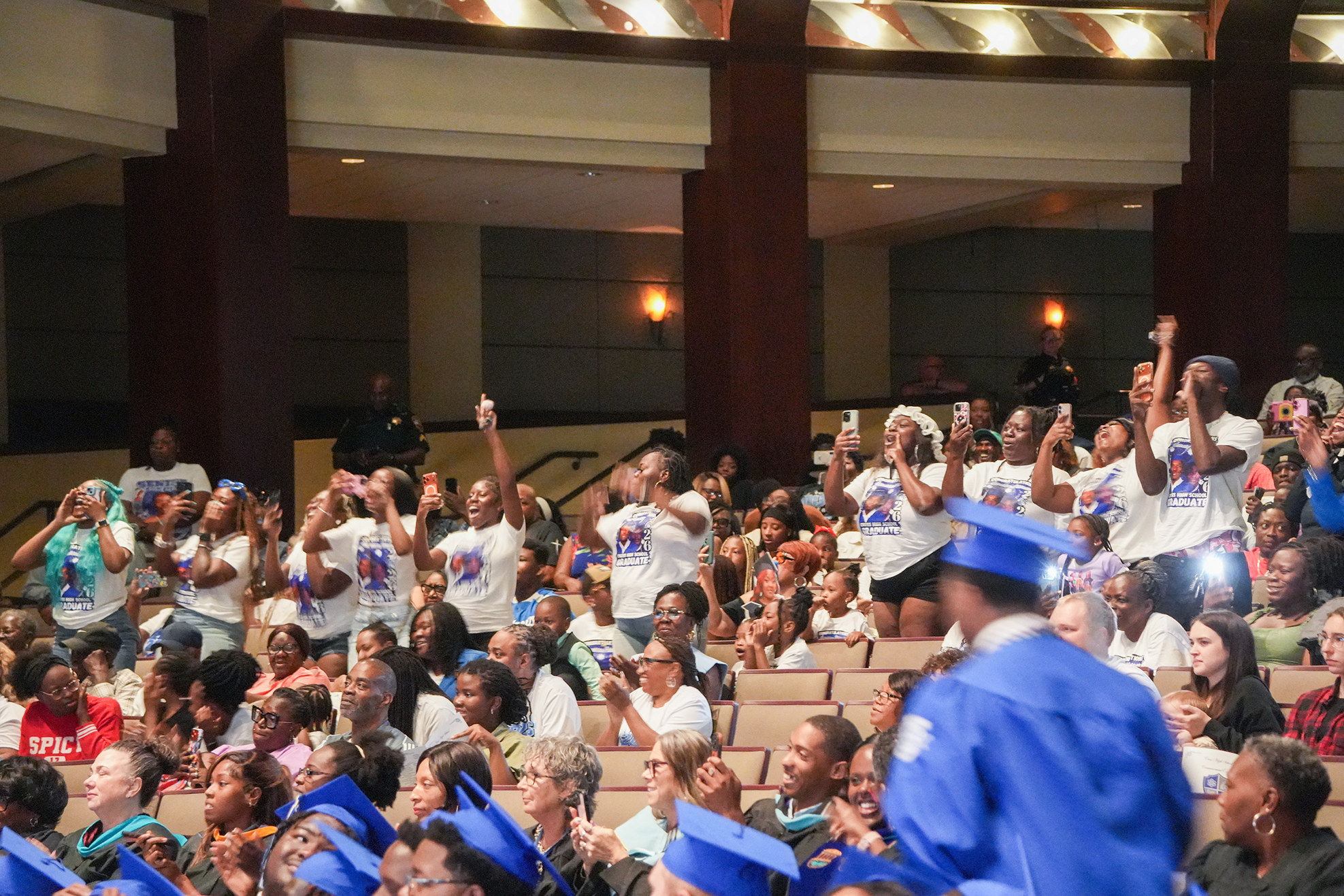 A large group of people in white shirts and blue graduation hats cheering and clapping in a theater.