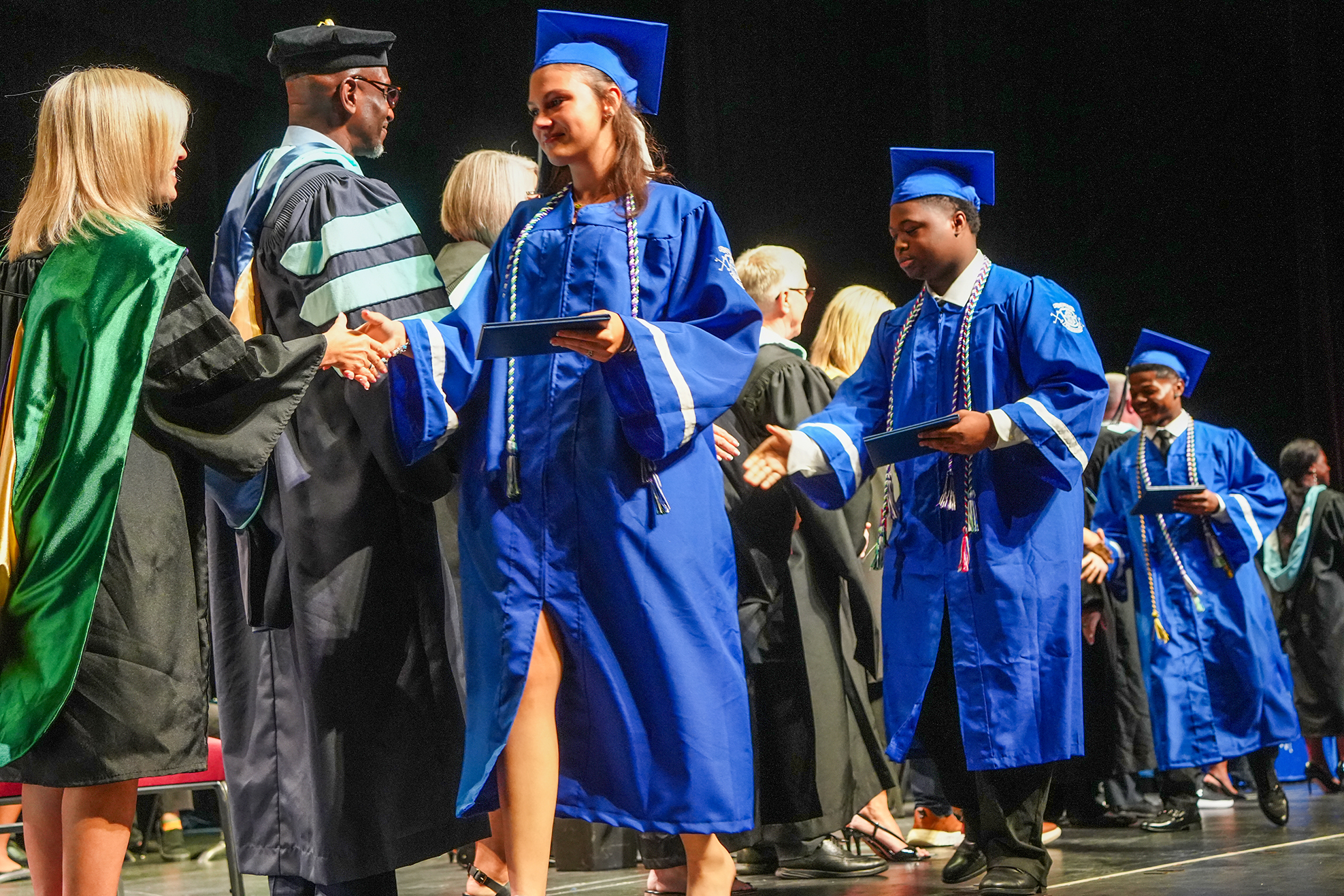 Graduates in blue gowns and mortarboards line up on stage, shaking hands with faculty. A person in green and black gown stands at the forefront.