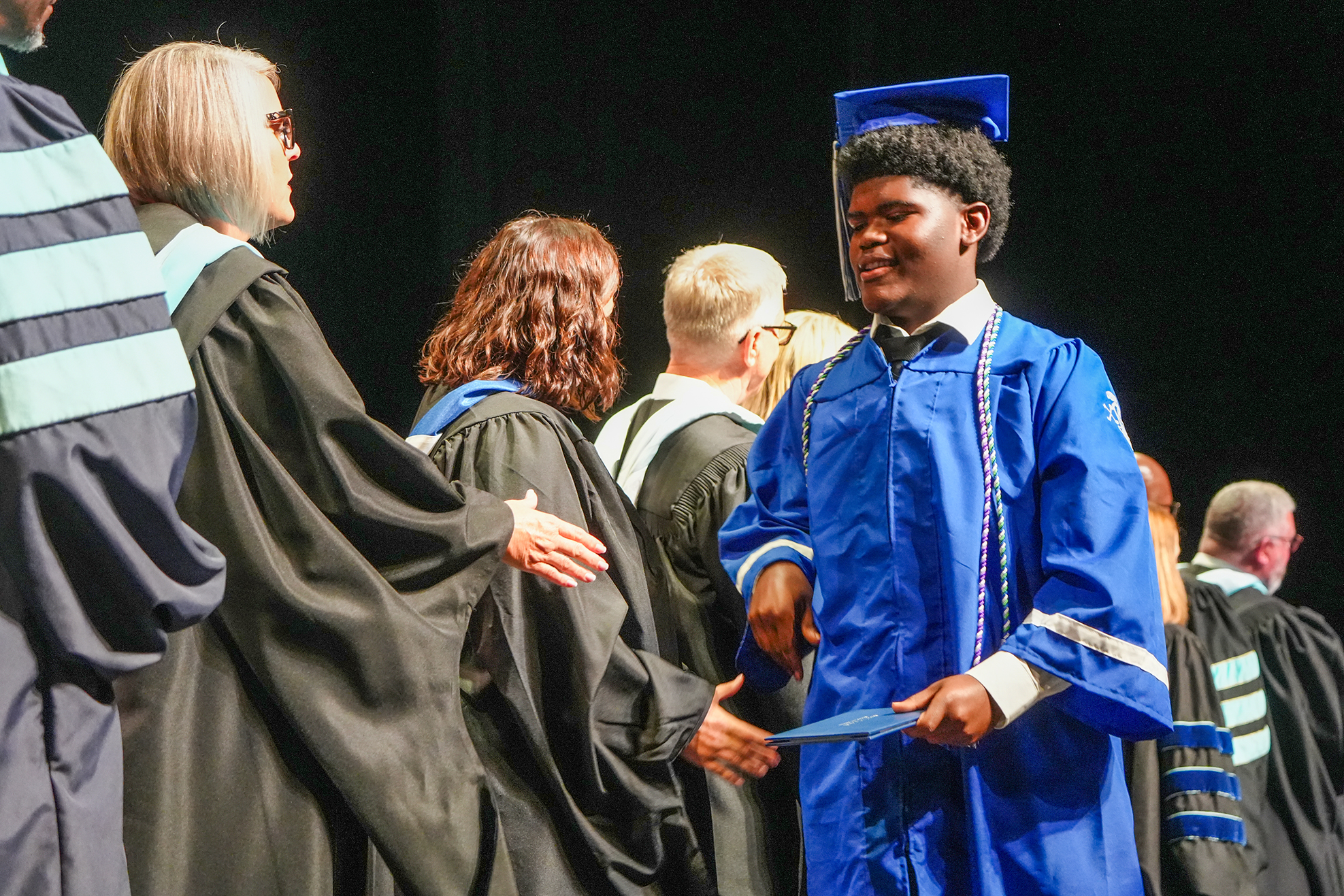 A young person in a blue graduation gown and cap receives a diploma from a woman in a black gown.